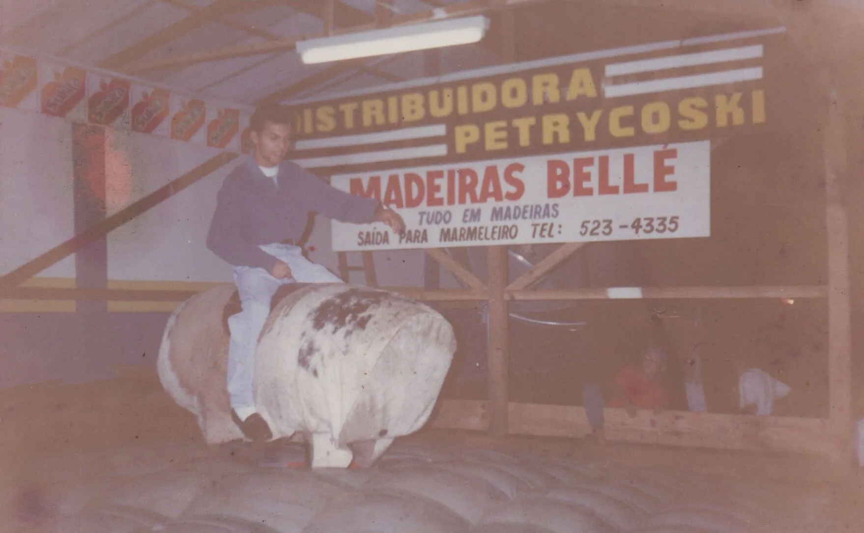Um homem de camisa azul e calças jeans está montado em um touro mecânico em um evento interno. Um banner com o texto "DISTRIBUIDORA PETRYKOSKI MADEIRAS BELLE" aparece atrás dele. (Original)