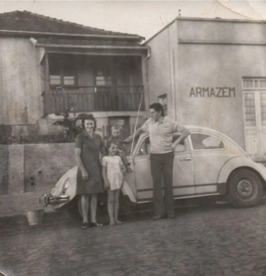 Família de quatro pessoas, incluindo dois adultos e duas crianças, posa ao lado de um Fusca bege vintage estacionado em uma rua de paralelepípedos na frente de edifícios antigos. (Original)
