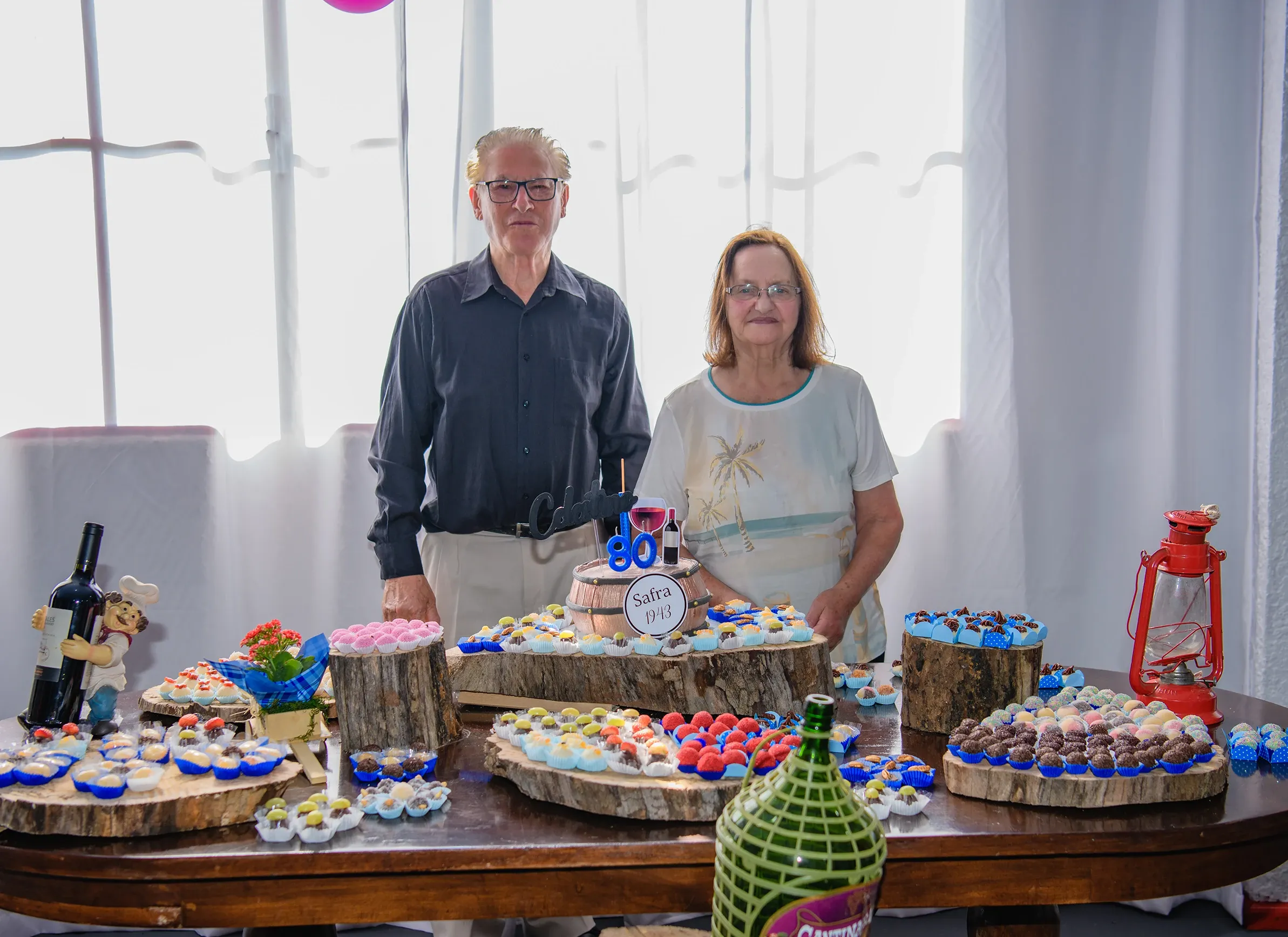 Um casal idoso, ele de camisa escura e ela de blusa clara, em pé atrás de uma mesa de aniversário com doces e um bolo de 80 anos.