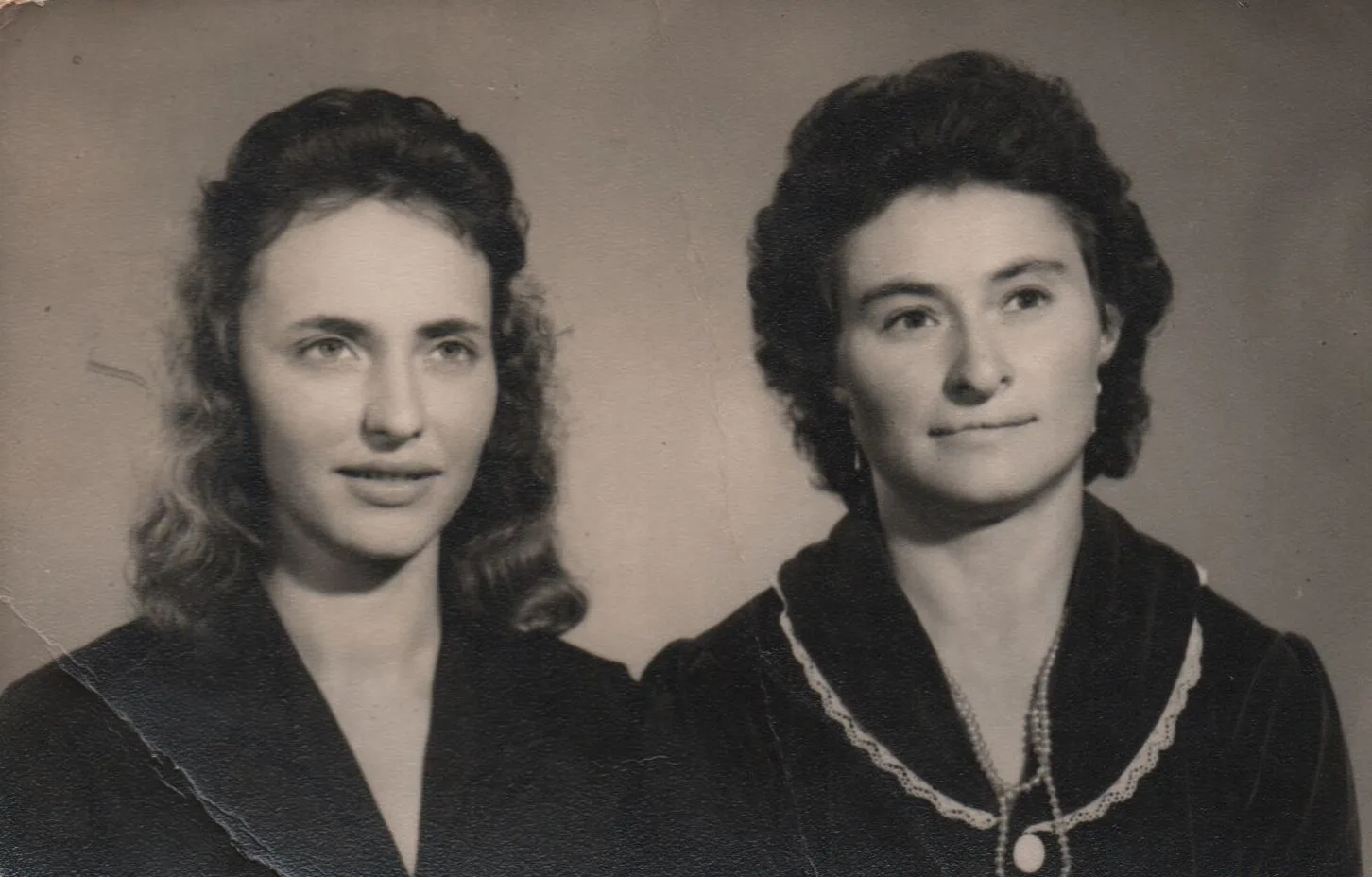 Retrato frontal de duas mulheres com pele clara. A mulher da esquerda tem cabelo castanho-claro encaracolado e usa um vestido azul-marinho. A mulher da direita tem cabelo castanho-escuro ondulado e usa um vestido cinza com um colar de pérolas. (Original)