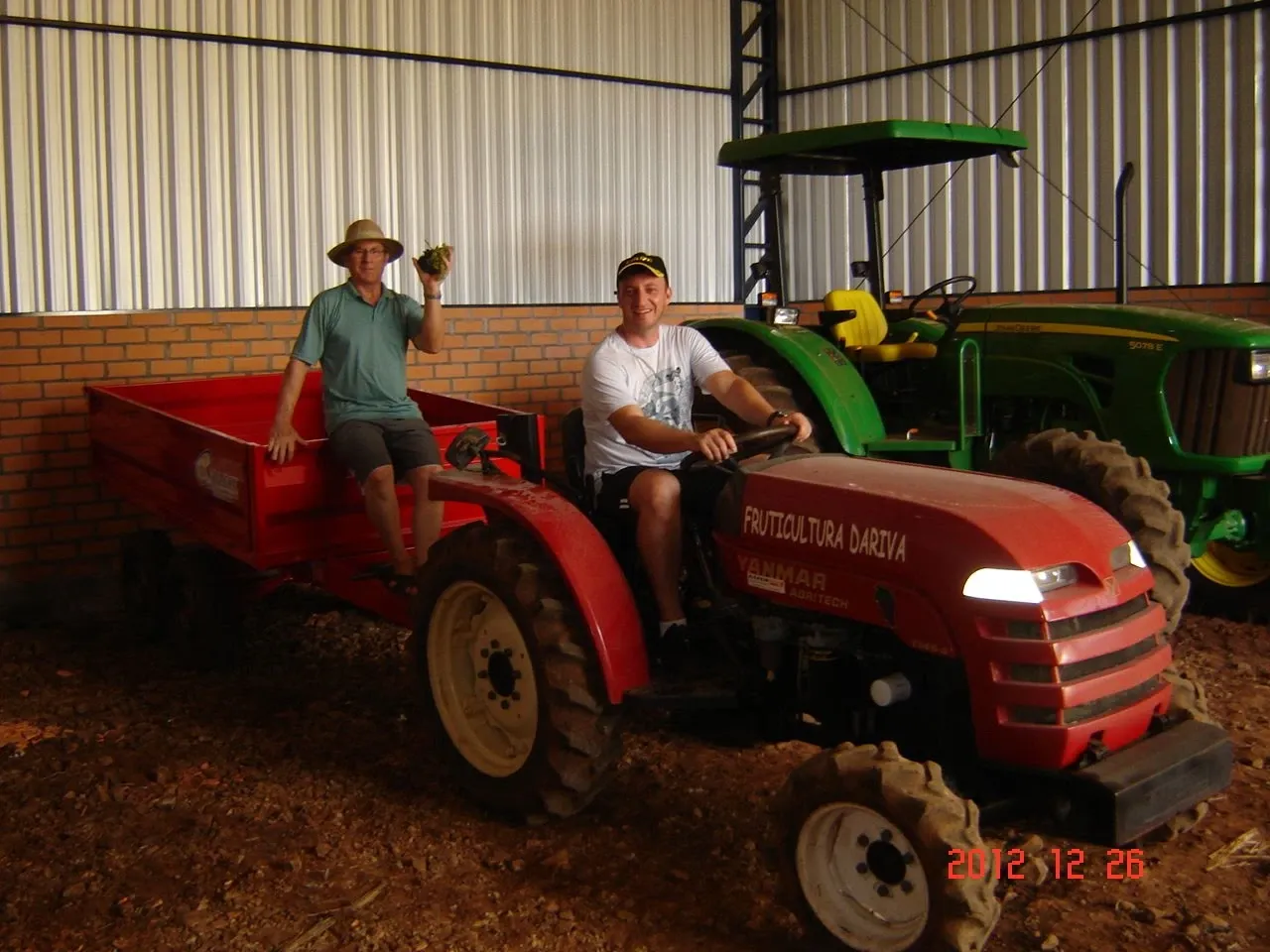Dois homens em um galpão. Um usa um chapéu de palha e segura uma fruta, sentado em um reboque vermelho acoplado a um trator vermelho. O outro sorri enquanto dirige o trator. Um segundo trator verde é visível no fundo.