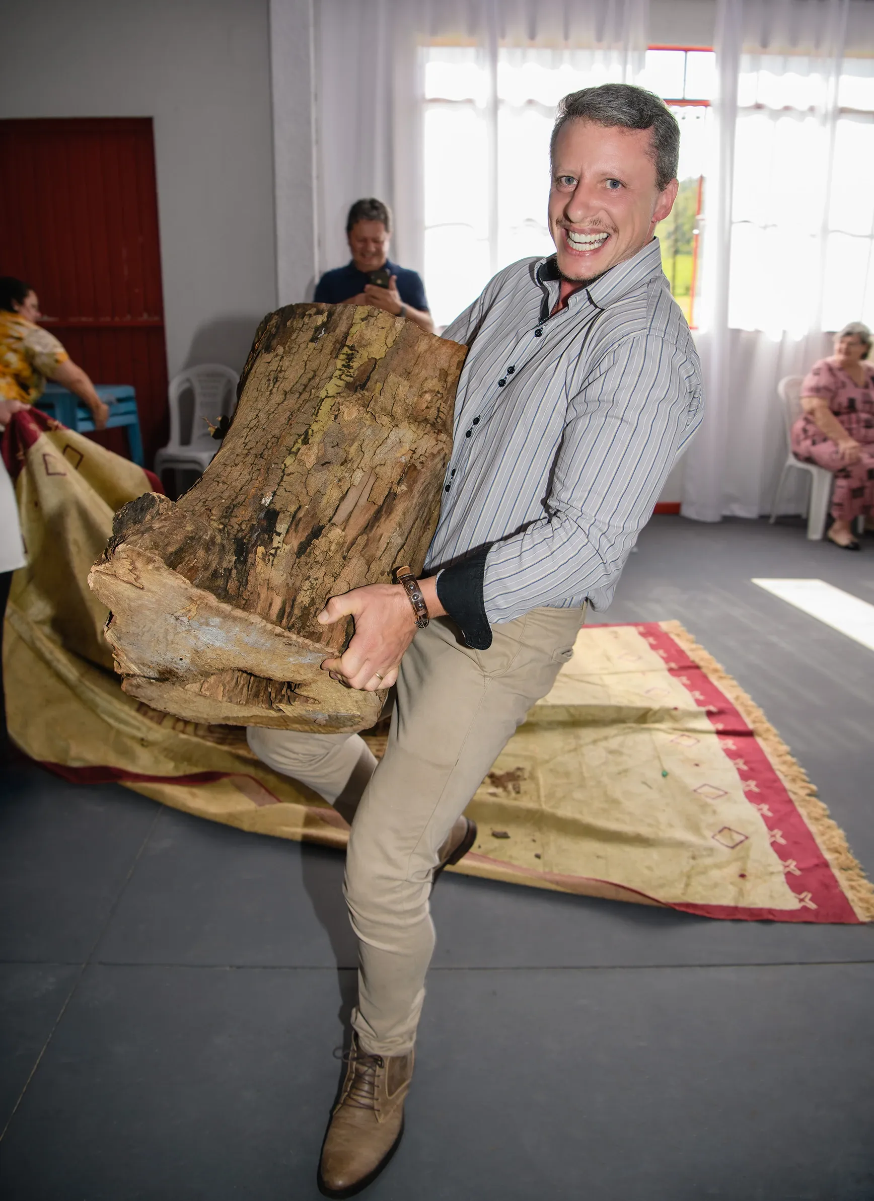 Um homem de meia-idade e cabelos grisalhos, vestindo uma camisa de botão listrada, calças cáqui e sapatos marrons, sorri para a câmera enquanto levanta um grande tronco de árvore sobre um tapete estampado, com outras pessoas em segundo plano.