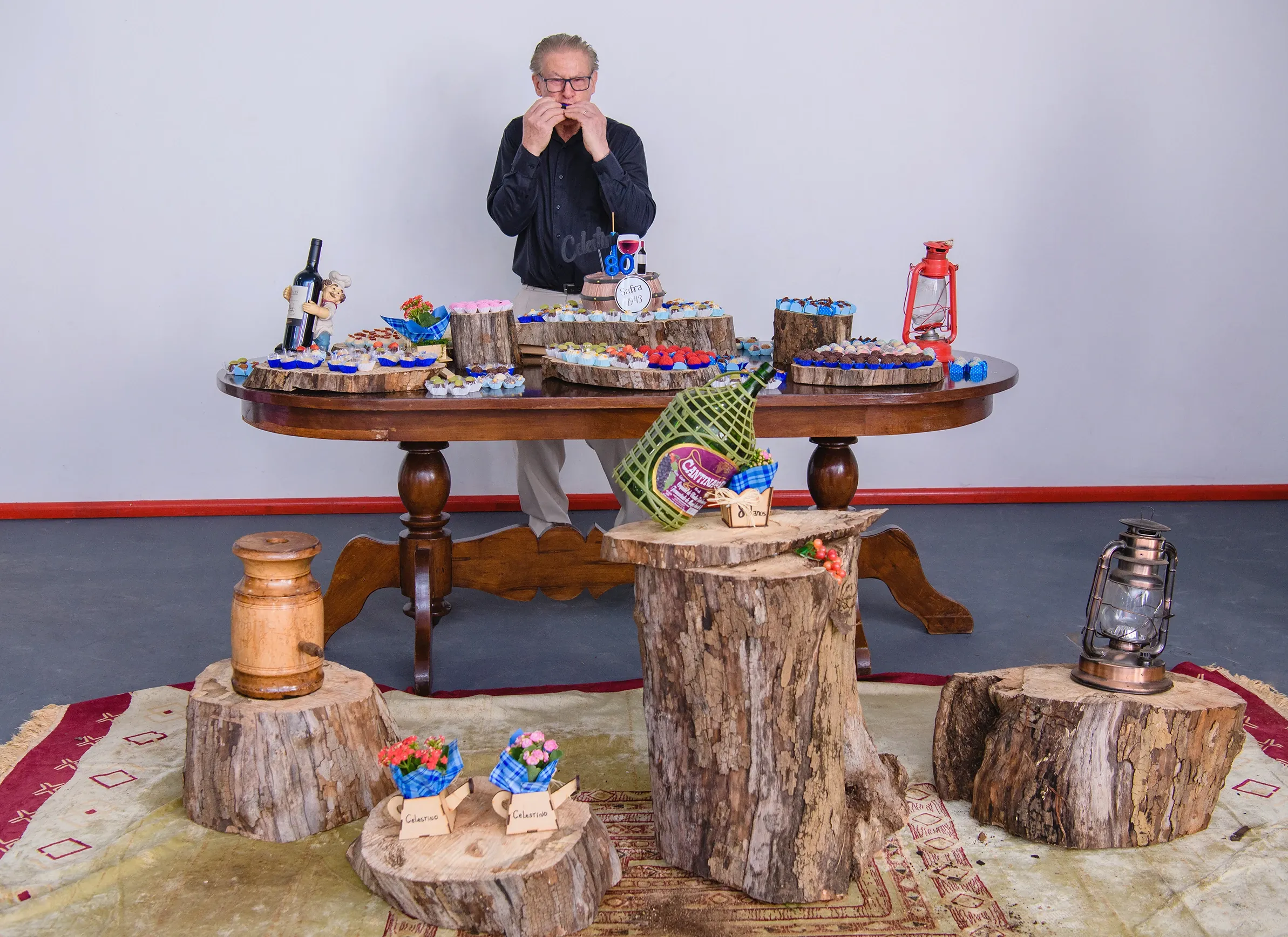 Um homem de camisa escura e calças claras está de pé atrás de uma mesa de madeira decorada com diversos doces, vinhos e um bolo, aparentemente em uma festa; o ambiente tem um fundo branco simples e alguns tocos de madeira com arranjos florais no chão.