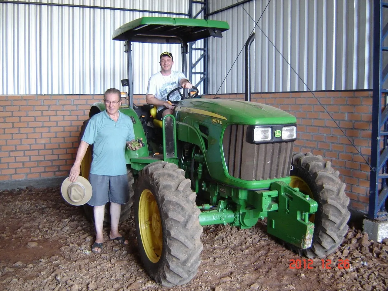 Dois homens perto de um trator verde. Um homem está sentado no banco do motorista e o outro está em pé no chão.