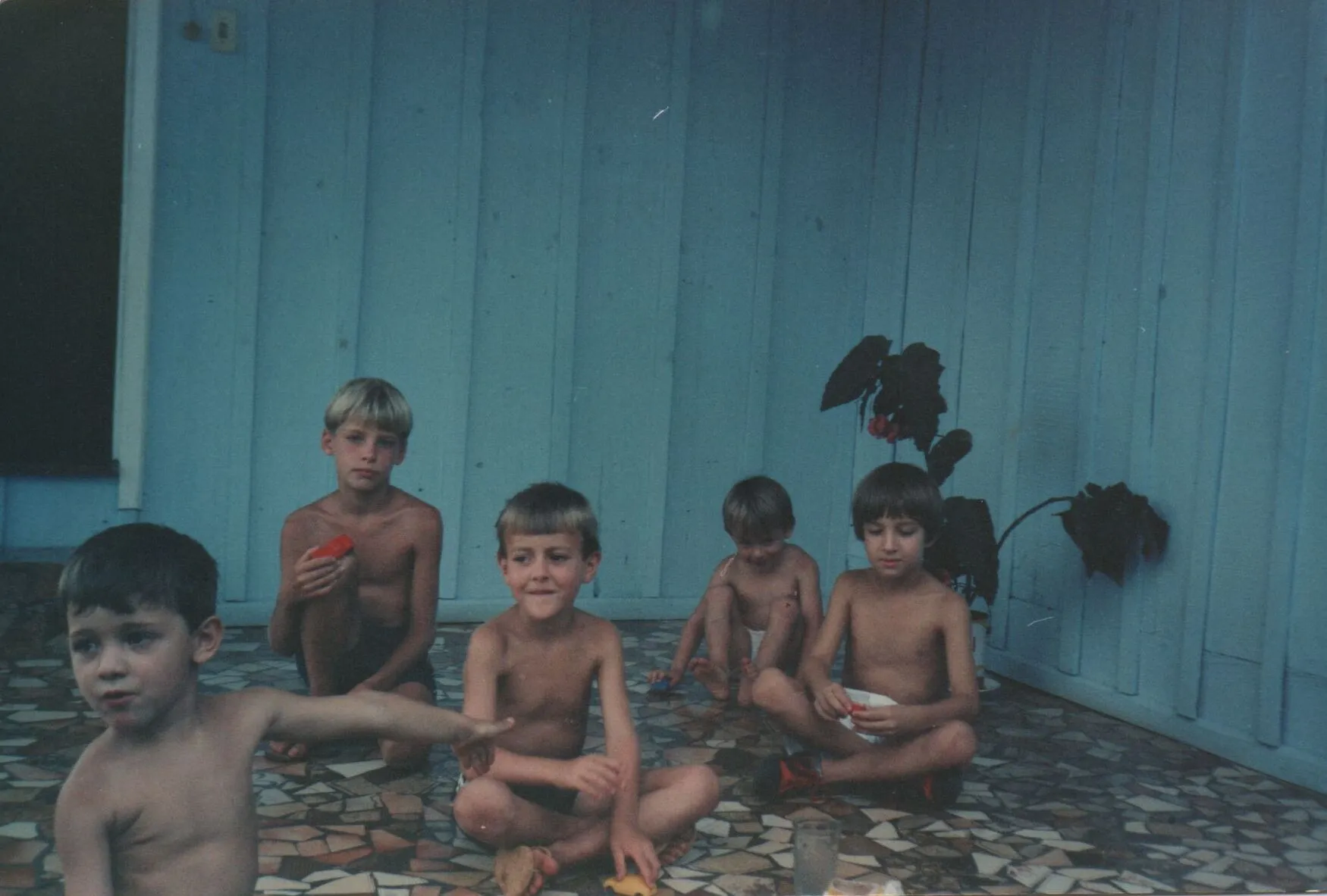 Cinco meninos sem camisa de 5 a 10 anos sentados em um pátio de azulejos marrons na frente de uma parede azul, alguns com as pernas cruzadas. (Original)