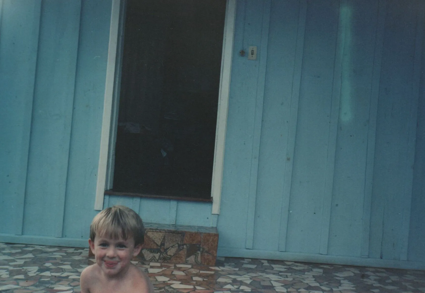 Um menino de pele clara e cabelo loiro curto está sem camisa e molhado, sorrindo diretamente para a câmera, com o que parece ser leite em seu lábio superior. Ele está em primeiro plano, sentado sobre um piso de pátio texturizado em tons terrosos, com uma parede azul de madeira e uma porta escura ao fundo. (Original)