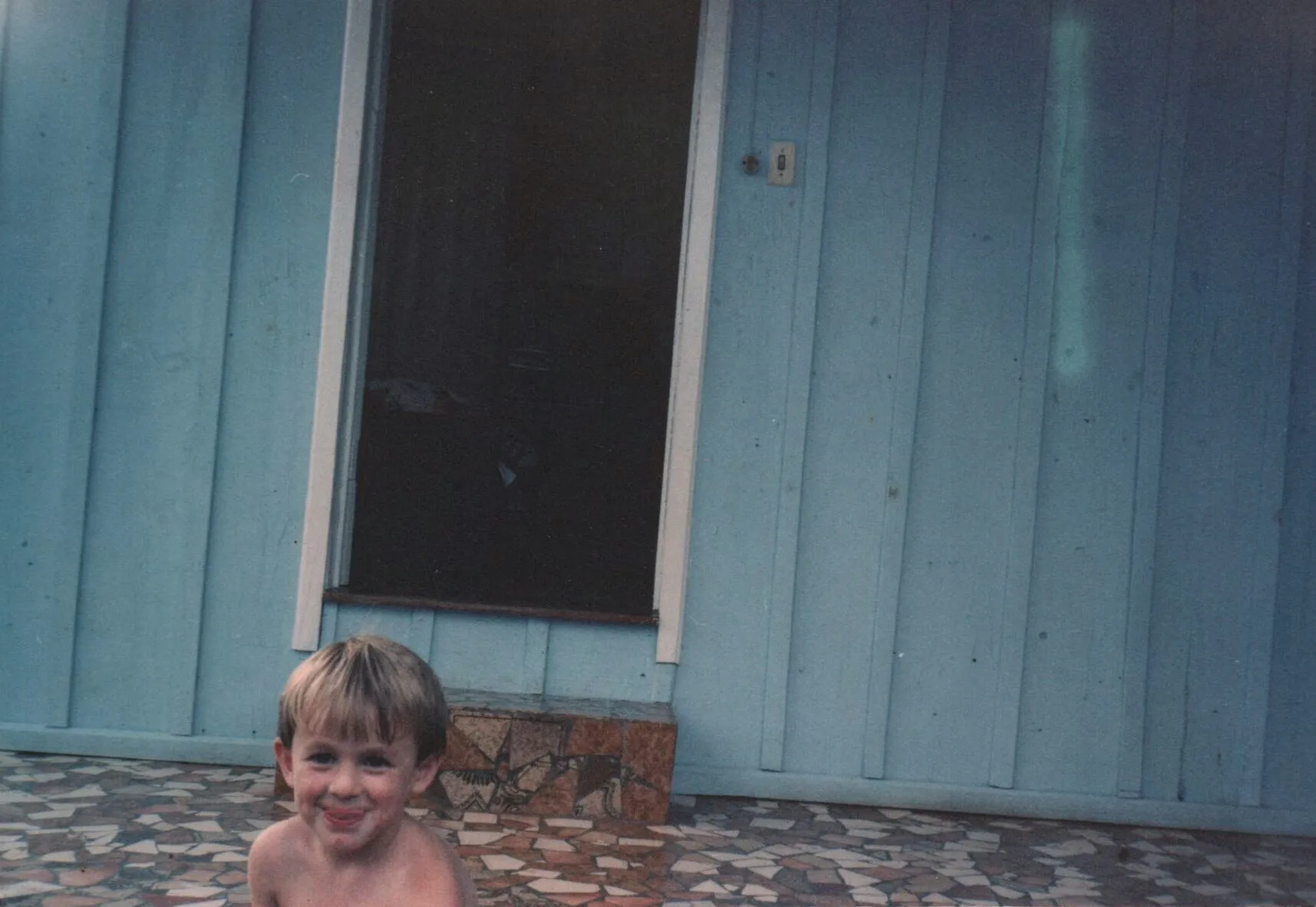 Um menino de pele clara e cabelo loiro curto está sem camisa e molhado, sorrindo diretamente para a câmera, com o que parece ser leite em seu lábio superior. Ele está em primeiro plano, sentado sobre um piso de pátio texturizado em tons terrosos, com uma parede azul de madeira e uma porta escura ao fundo.