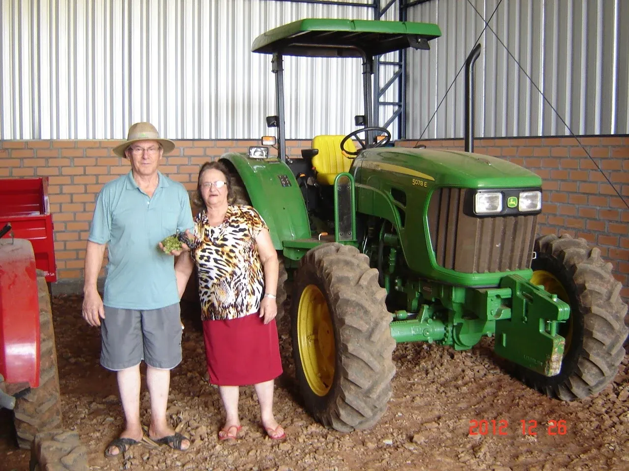 Um casal idoso, com a mulher vestindo uma saia vermelha e uma blusa estampada, e o homem com um chapéu e camisa polo azul, está em pé na frente de um trator verde em um galpão.