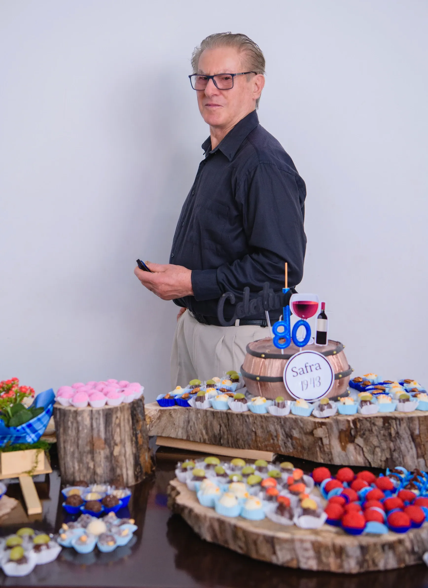 Homem mais velho de óculos e camisa preta em pé ao lado de uma mesa com bolo e doces.