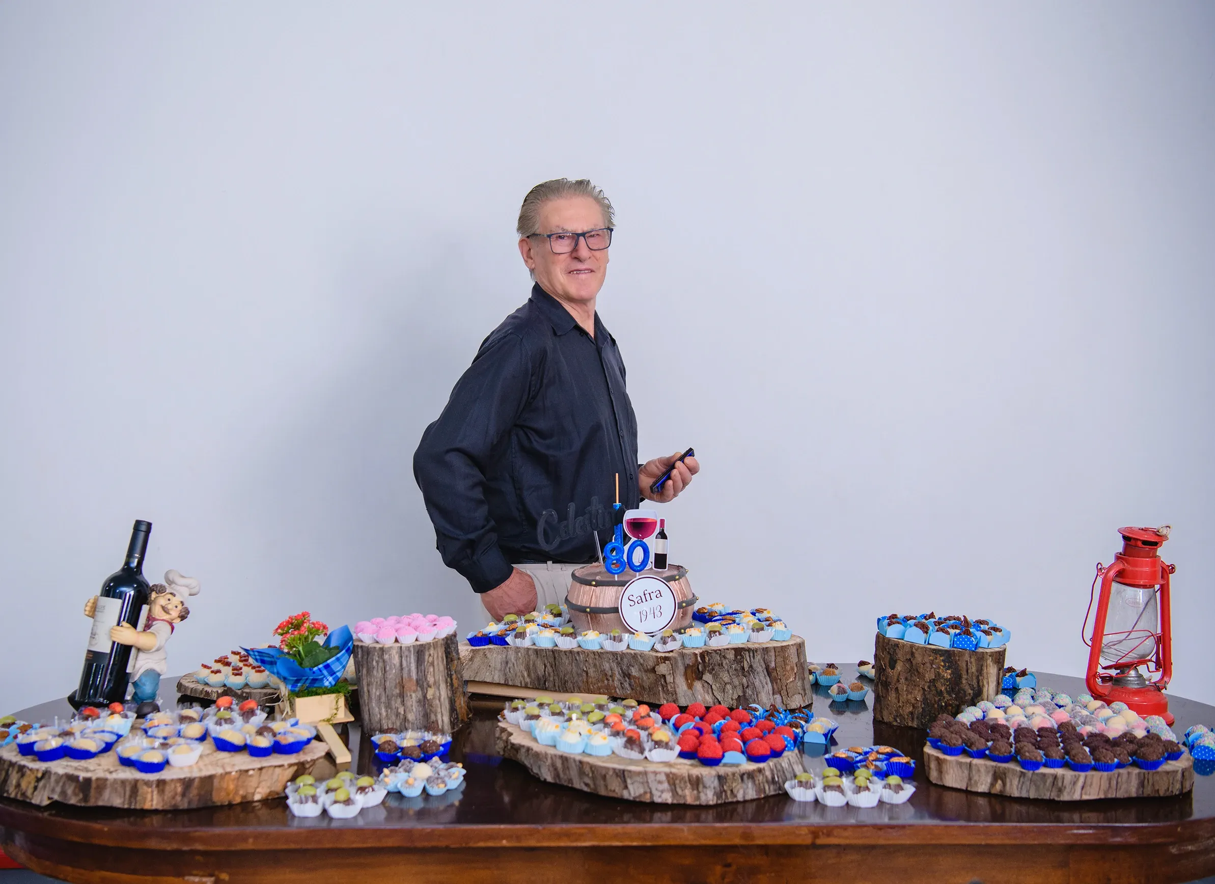 Um homem de camisa preta e óculos está atrás de uma mesa de sobremesas de festa, olhando por cima do ombro para a câmera, com um bolo de dois andares e petiscos dispostos em travessas de madeira.