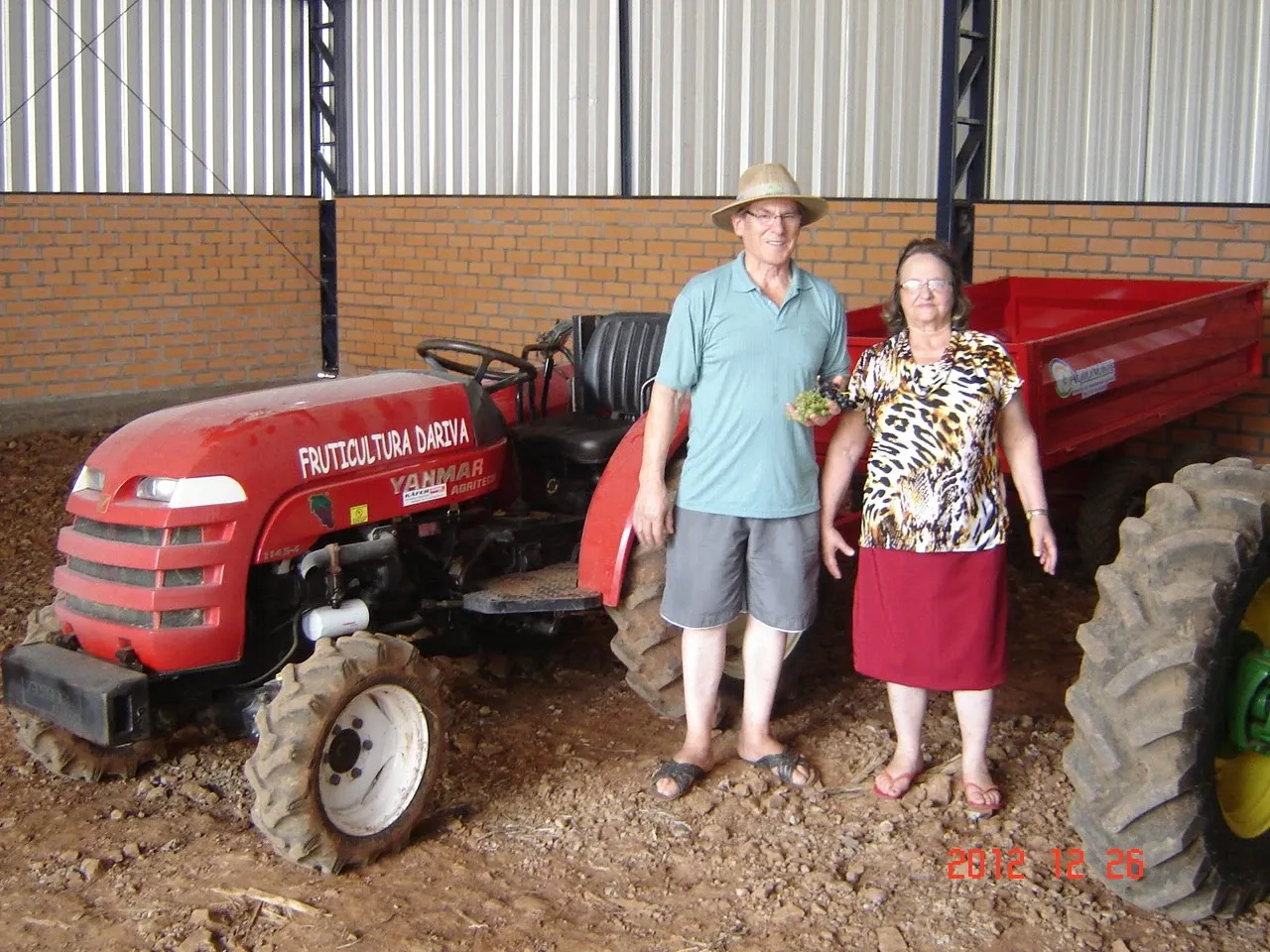 Um homem e uma mulher idosos posam ao lado de um trator vermelho em uma fazenda.