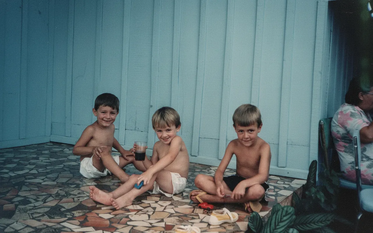 Três meninos sorridentes sentados em um chão de azulejos na varanda, um deles segurando um copo, com uma mulher sentada em uma cadeira ao fundo à direita.