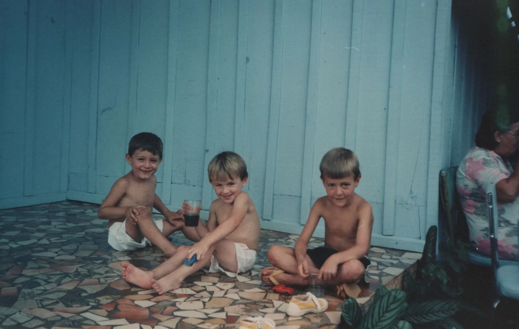 Três meninos sorridentes sentados em um chão de azulejos na varanda, um deles segurando um copo, com uma mulher sentada em uma cadeira ao fundo à direita. (Original)