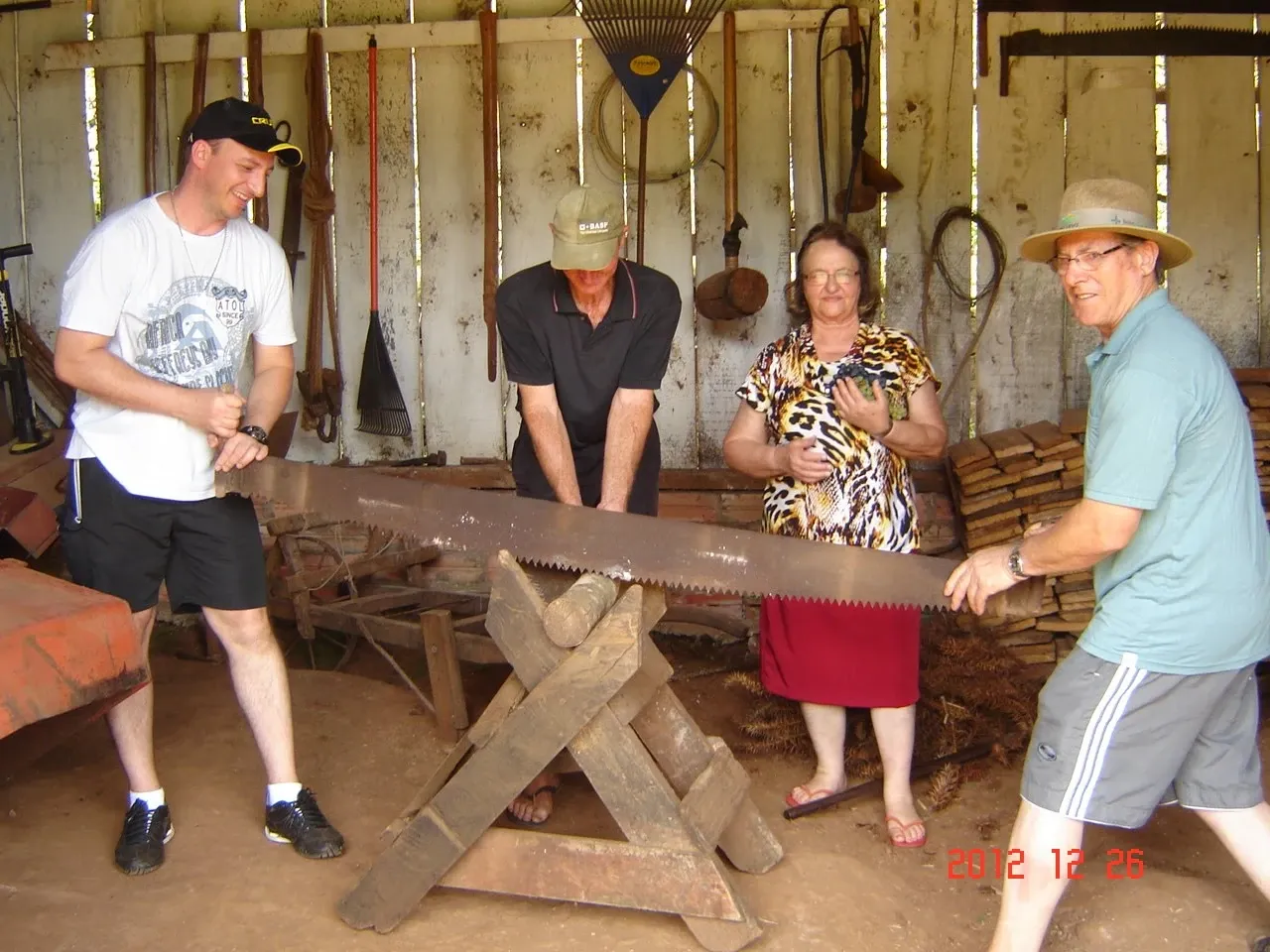 Quatro pessoas, dois homens e uma mulher, serram uma grande tora com um serrote em uma oficina rústica cheia de ferramentas.