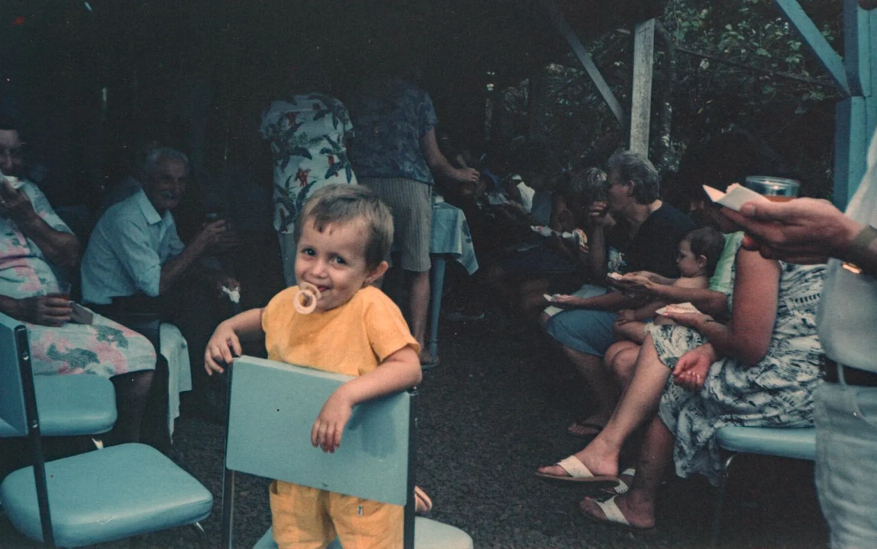 Uma criança pequena com uma chupeta na boca está sorrindo para a câmera, segurando as laterais de uma pequena cadeira azul em uma reunião de família ao ar livre.