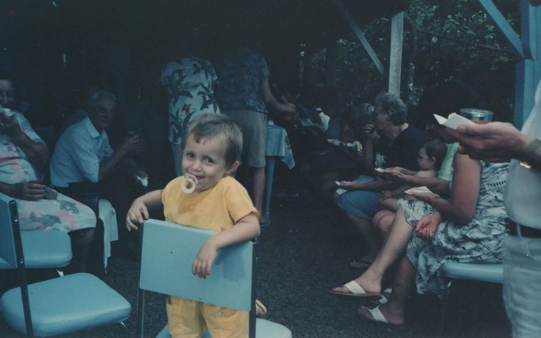 Uma criança pequena com uma chupeta na boca está sorrindo para a câmera, segurando as laterais de uma pequena cadeira azul em uma reunião de família ao ar livre. (Original)
