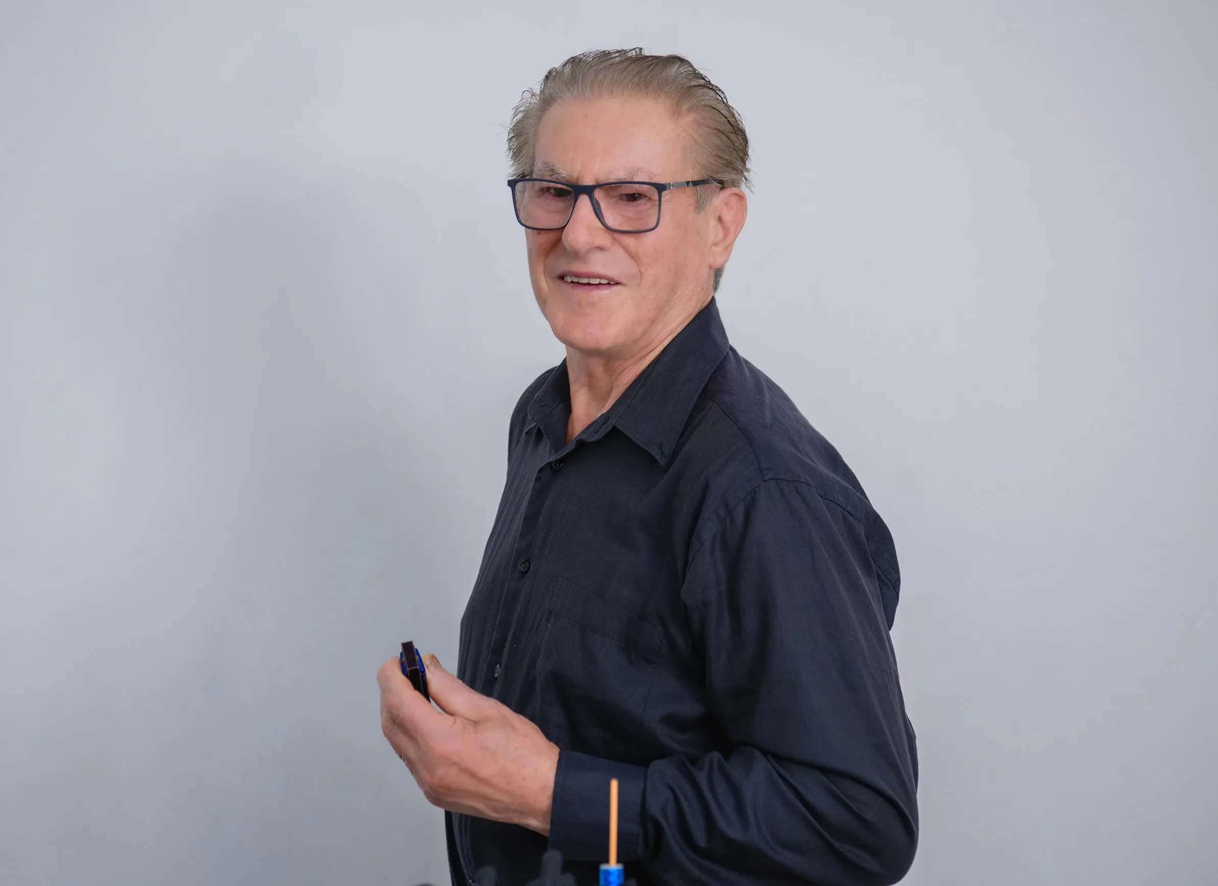 Homem idoso de óculos, camisa social escura, sorrindo e segurando uma caneta, em frente a uma parede branca.