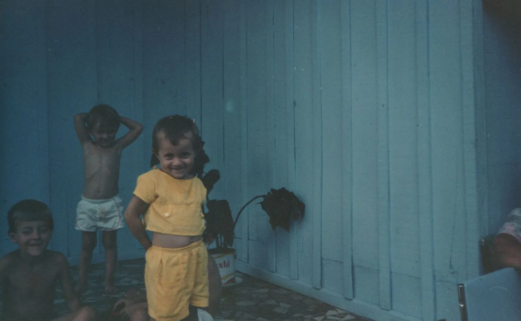 Três crianças com tons de pele claros e cabelos castanhos curtos brincam no que parece ser uma varanda. A criança à esquerda está sorrindo enquanto se senta no chão sem camisa. A criança no meio está em pé sem camisa, com as mãos atrás da cabeça, e a criança à direita está em pé sorrindo, vestindo uma camisa amarela e shorts amarelos. Uma parede azul e plantas em vasos podem ser vistas ao fundo. (Original)
