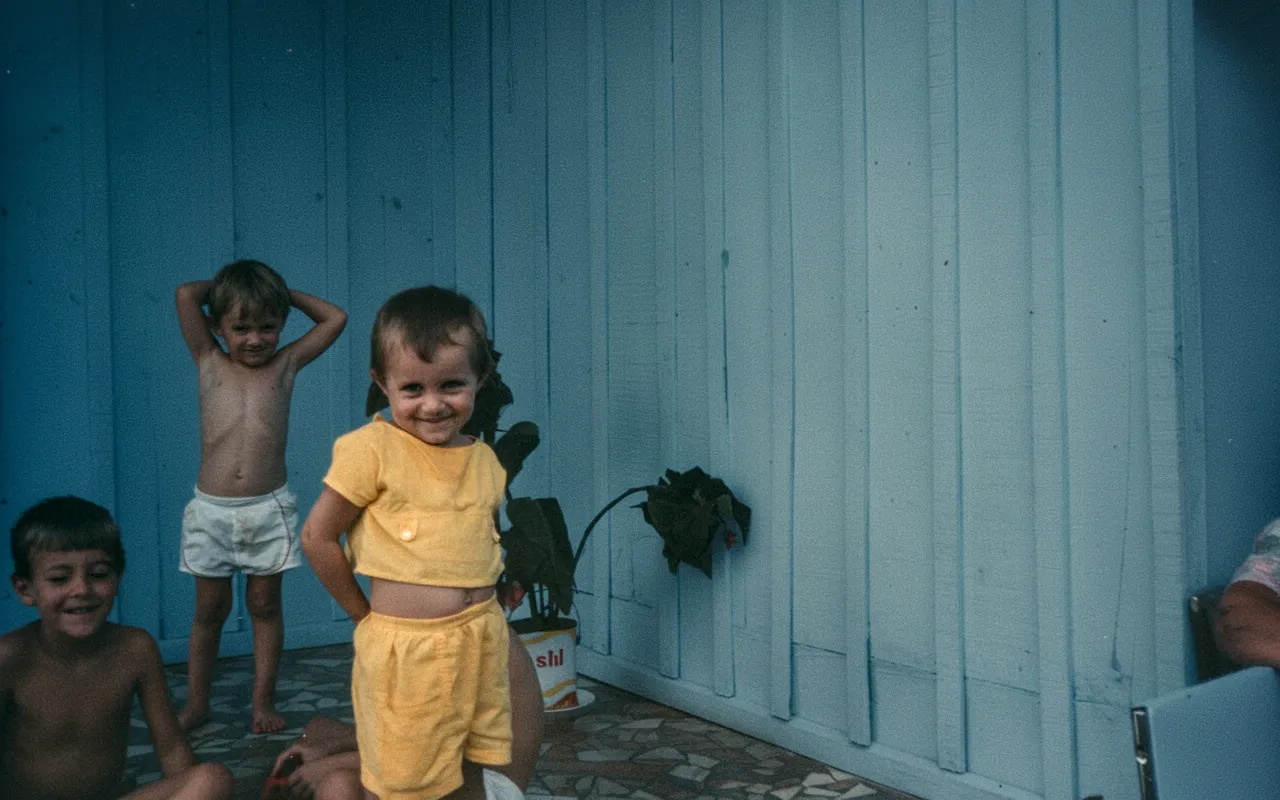 Três crianças com tons de pele claros e cabelos castanhos curtos brincam no que parece ser uma varanda. A criança à esquerda está sorrindo enquanto se senta no chão sem camisa. A criança no meio está em pé sem camisa, com as mãos atrás da cabeça, e a criança à direita está em pé sorrindo, vestindo uma camisa amarela e shorts amarelos. Uma parede azul e plantas em vasos podem ser vistas ao fundo.
