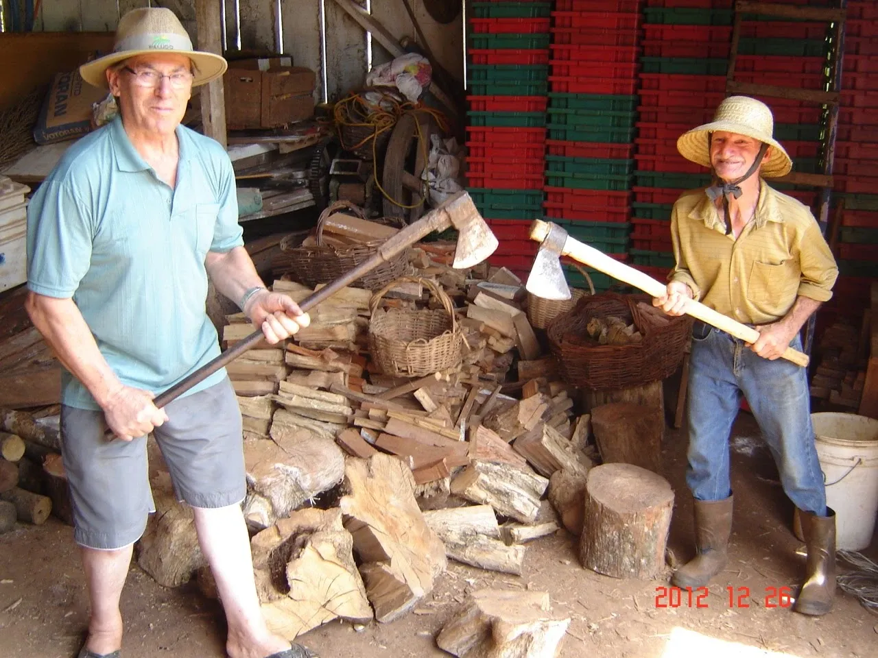 Dois homens sorridentes, usando chapéus de palha e segurando machados, estão em uma pilha de madeira na frente de uma parede de engradados coloridos e uma cabana rústica.