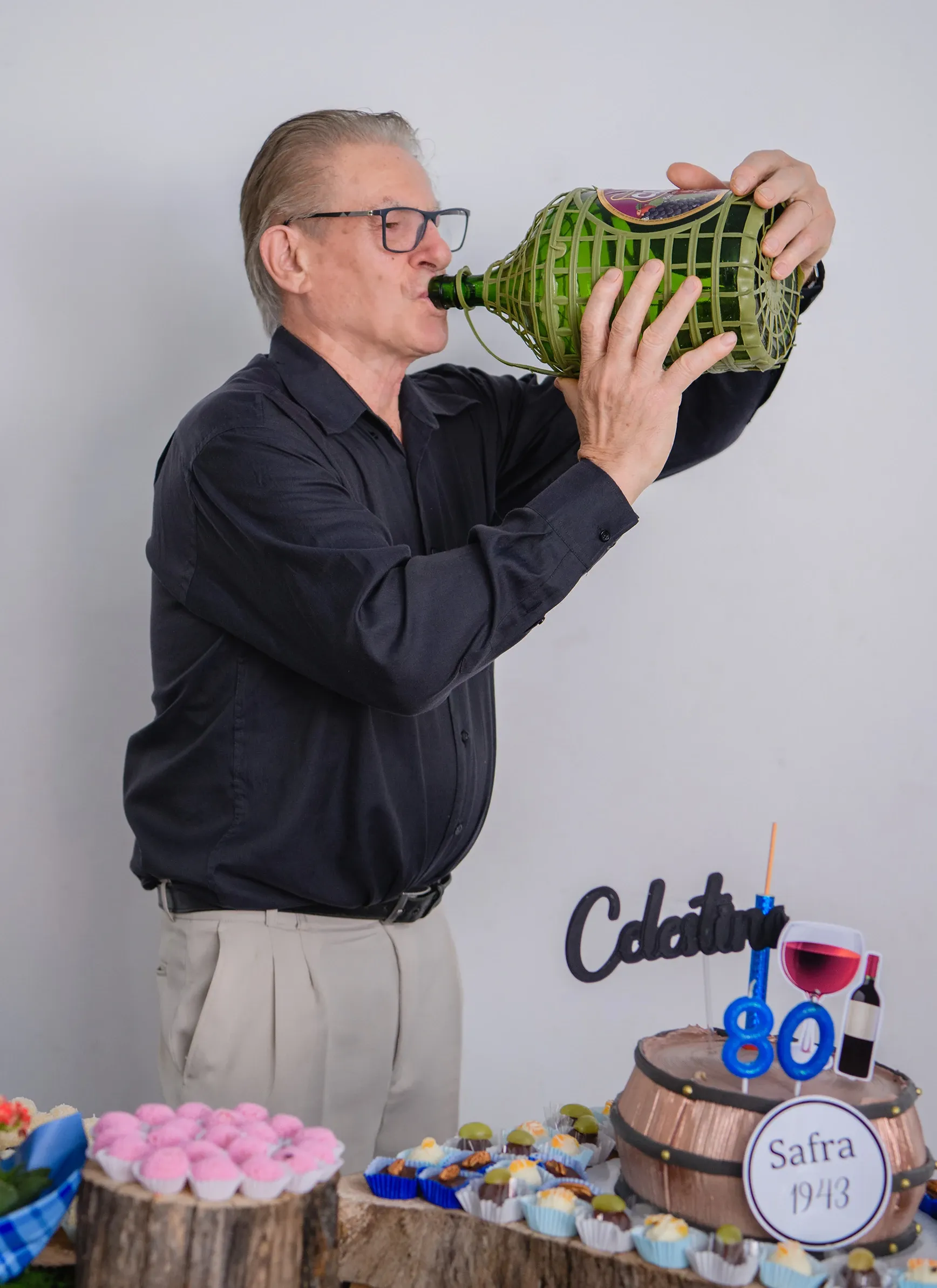 Um homem de meia-idade, com óculos e camisa preta, bebe diretamente de uma grande garrafa de vinho com cesta de palha em uma festa de aniversário de 80 anos com bolo de vinho e doces.