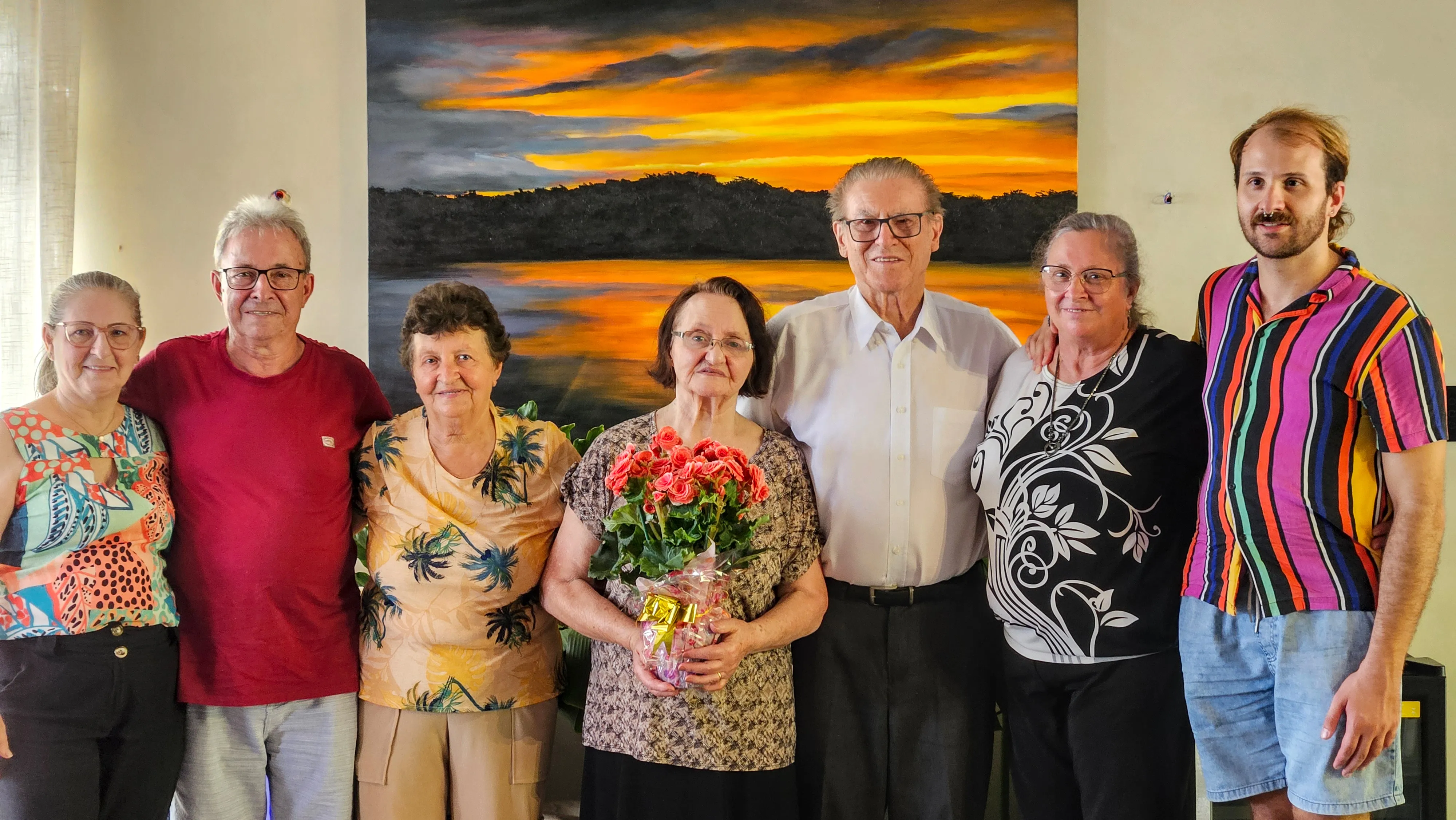 Um grupo de seis pessoas, duas das quais são idosas, posa para uma foto em frente a uma pintura paisagística.