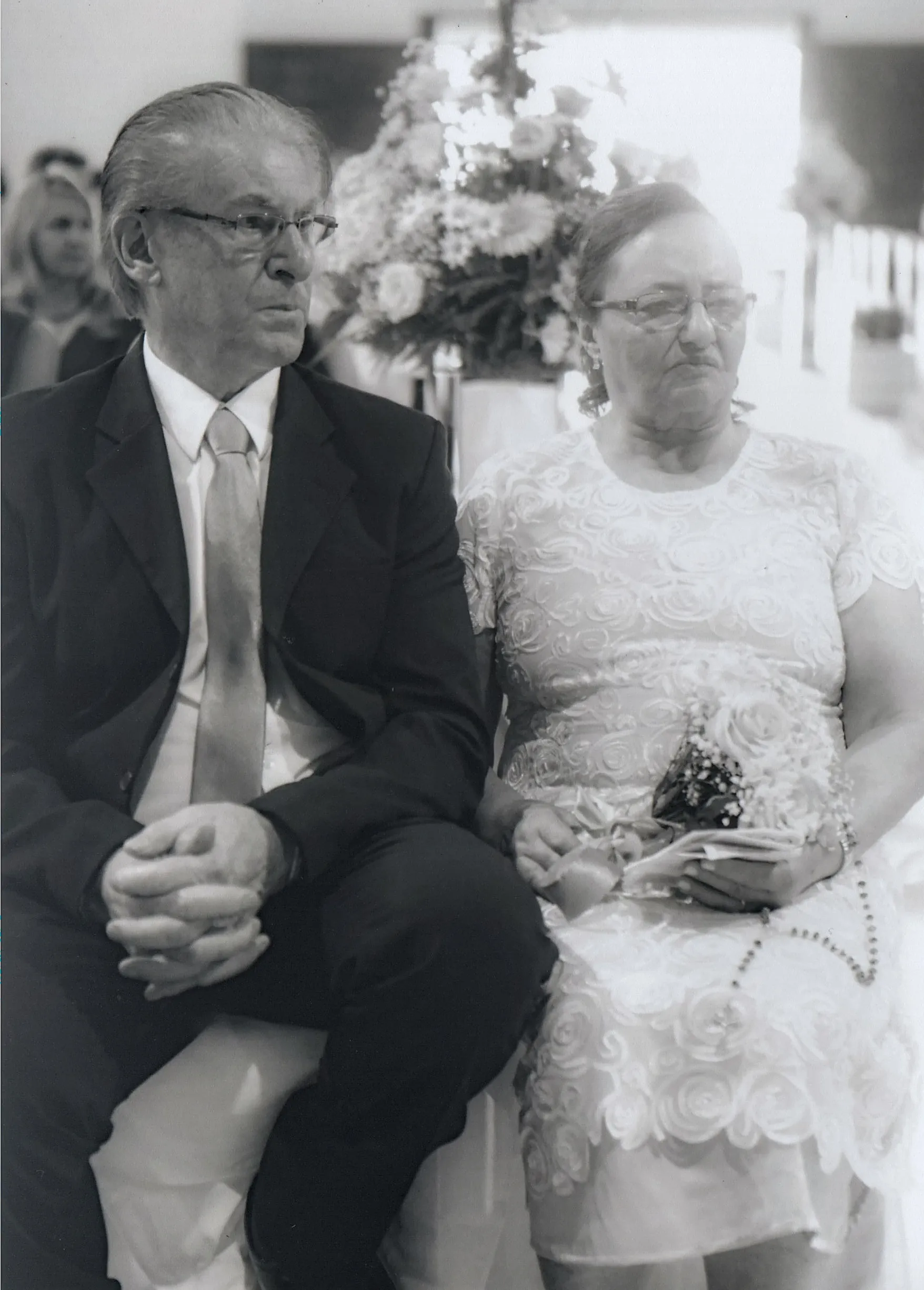 Um casal de idosos sentados em frente a arranjos de flores; o homem à esquerda usa óculos e um terno com gravata, e a mulher à direita usa óculos e um vestido de renda branca enquanto segura um buquê e um rosário.