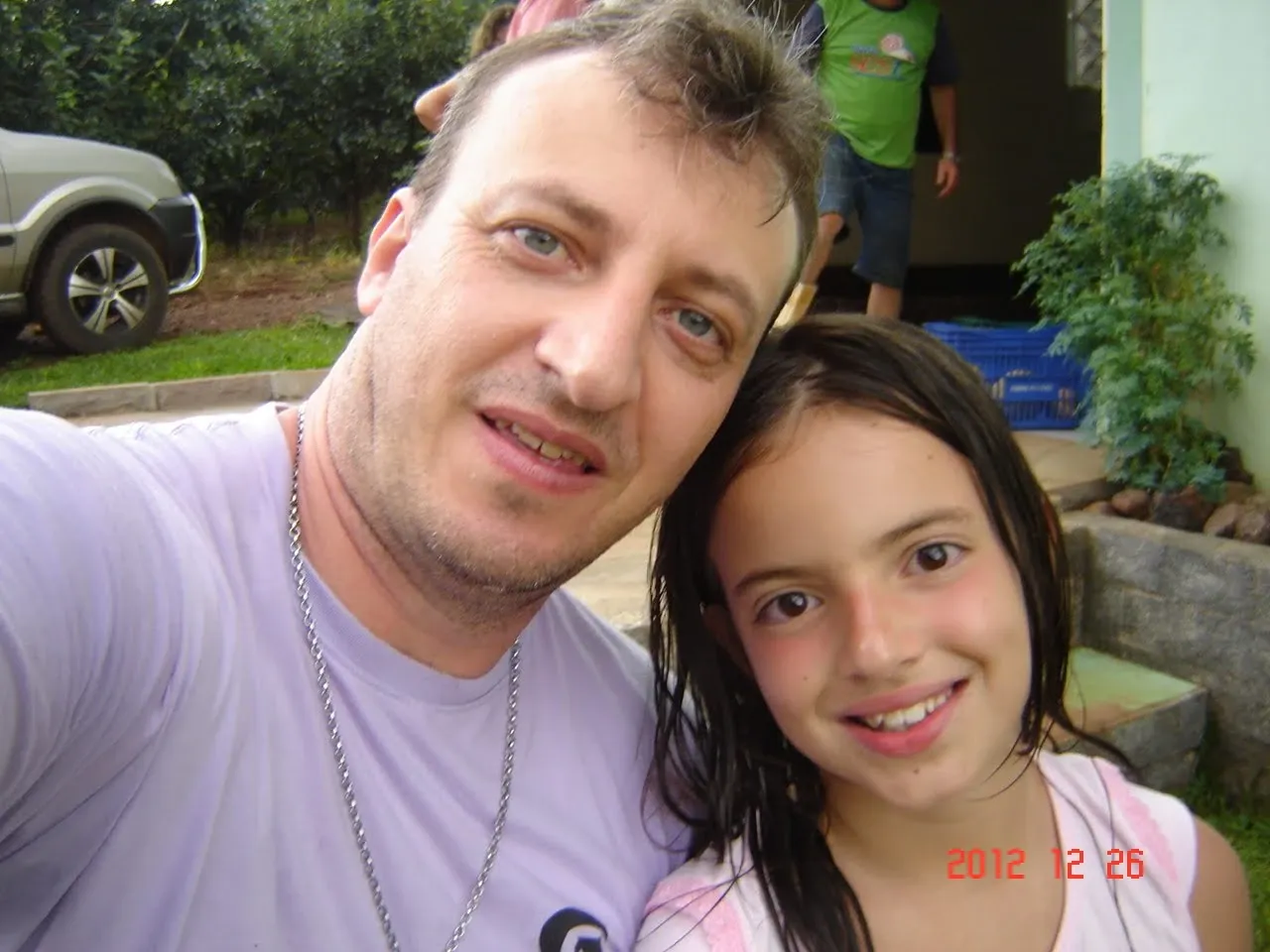 Homem de pele clara e cabelo castanho com uma corrente de prata sorri ao lado de uma menina de cabelo castanho comprido e molhado, ambos sorrindo para a câmera ao ar livre.