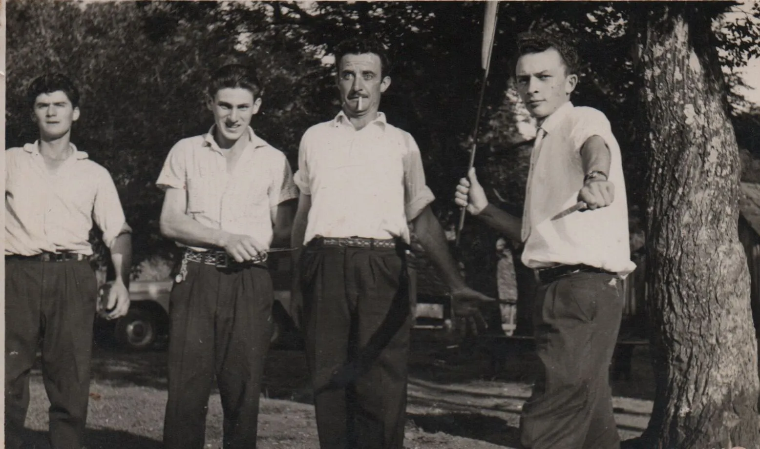 Quatro homens estão de pé e olhando para frente em um gramado sombrio, possivelmente um quintal, e três deles estão sorrindo. (Original)