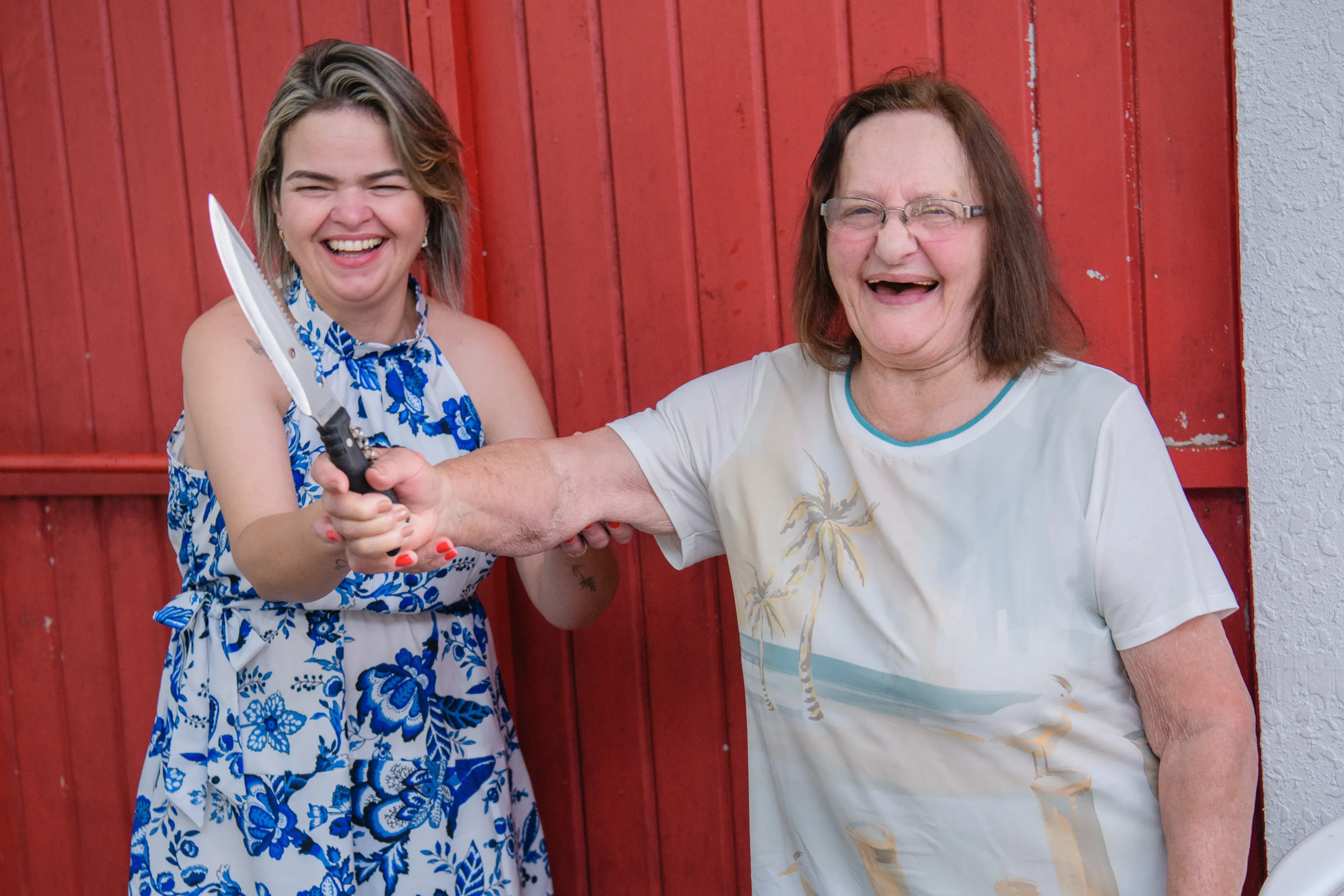 Duas mulheres, uma jovem e uma idosa, riem enquanto a jovem segura uma faca para a câmera.