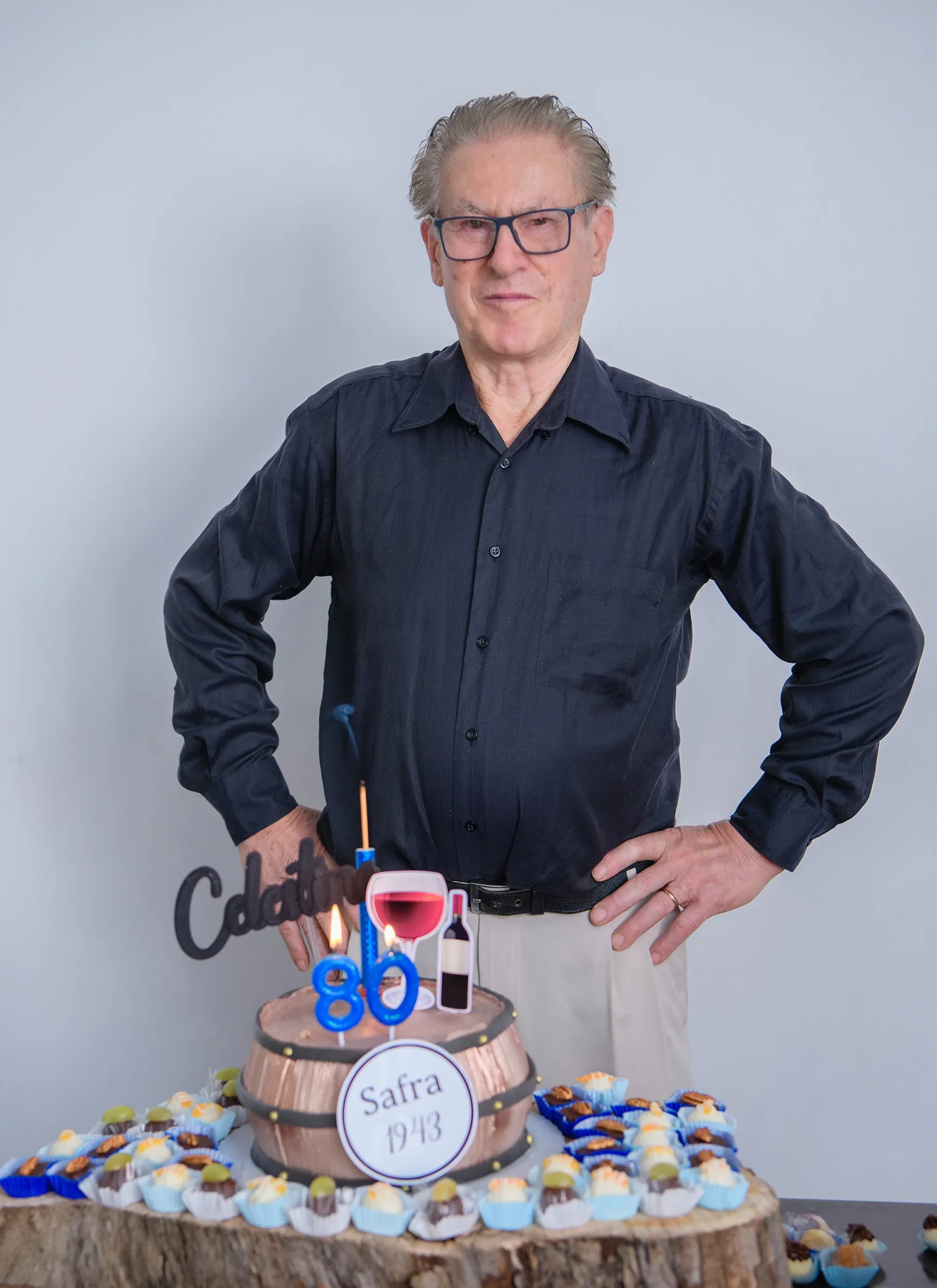 Homem de meia-idade sorrindo para a câmera, com as mãos nos quadris, atrás de um bolo de aniversário decorado com o número 80, ao lado de vários doces em uma mesa de madeira.