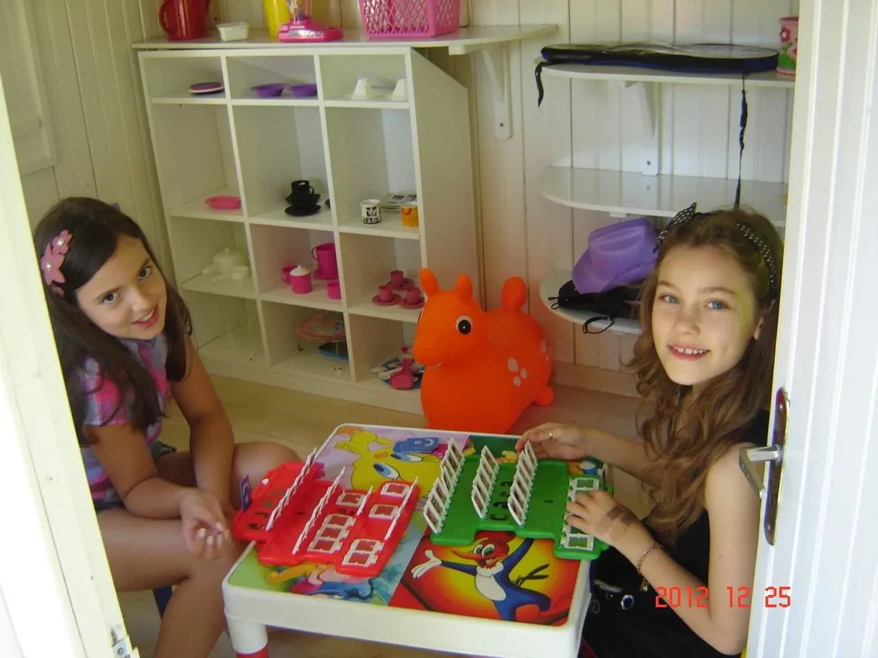 Duas meninas, uma sentada à esquerda em uma cadeira e outra à direita no chão, sorriem enquanto brincam com um jogo de tabuleiro em uma sala de brinquedos, com uma estante branca cheia de brinquedos rosa e brancos ao fundo e um brinquedo de cavalo laranja no chão.
