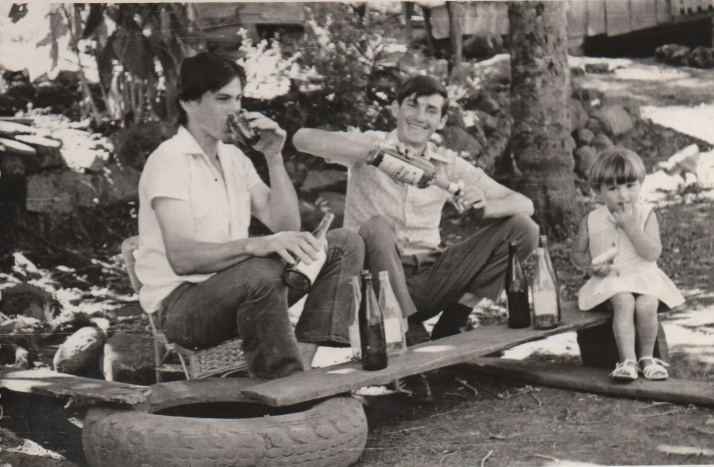 Dois homens bebem em um tábua improvisada com garrafas enquanto uma criança senta-se na ponta oposta. (Original)