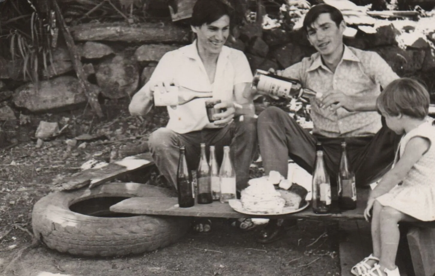 Dois homens sorridentes, sentados ao ar livre, servem bebidas de garrafas para copos e para outras garrafas, enquanto uma criança observa curiosamente ao lado de uma mesa improvisada com garrafas e comida. (Original)
