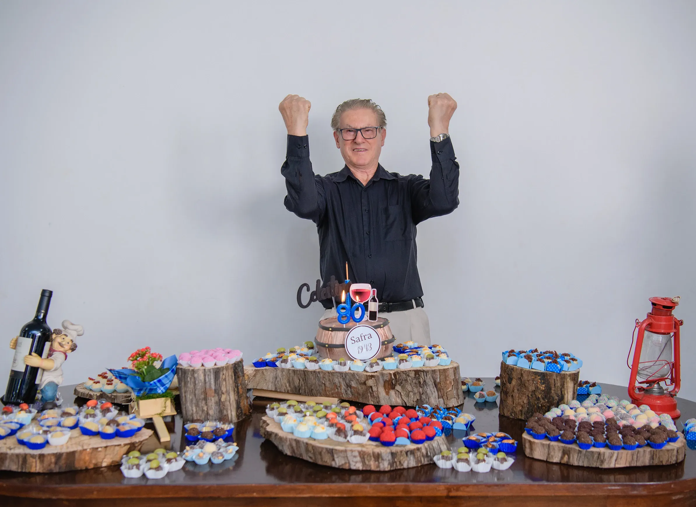 Um homem com óculos e camisa preta levanta os punhos em comemoração atrás de uma mesa cheia de doces e um bolo de aniversário com a frase "90 Safra 1933".