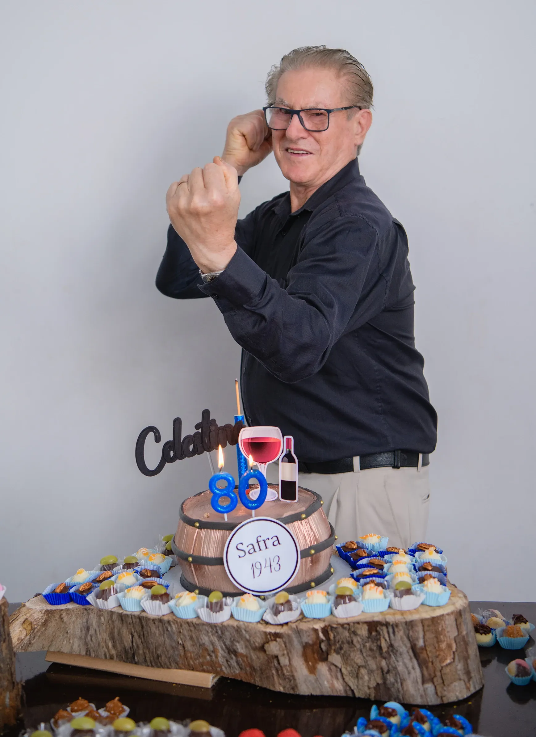 Um homem idoso com óculos e camisa escura está sorrindo, os punhos cerrados e um bolo de aniversário rústico de 80 anos com enfeites de vinho e doces em mini forminhas à sua frente, em um ambiente de celebração.