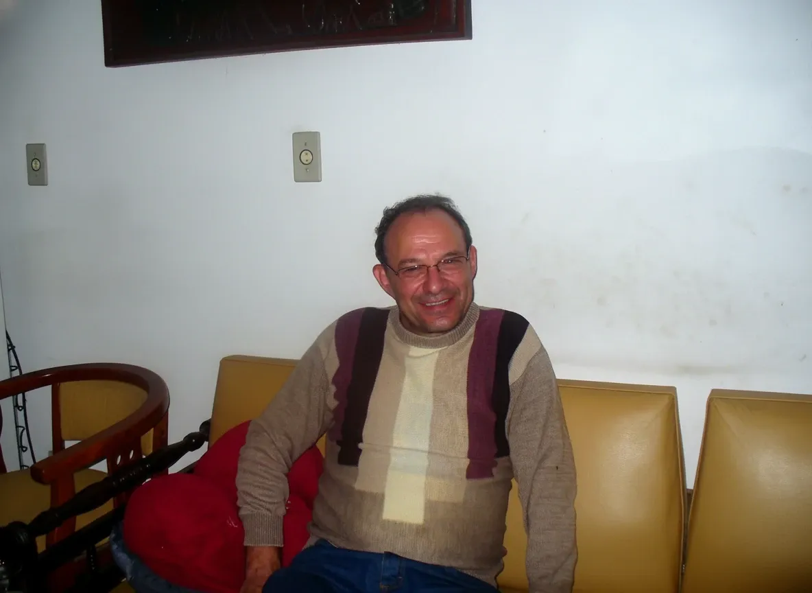 Homem sorridente de meia-idade com óculos, camisa listrada de malha e calça jeans azul, sentado em um sofá amarelo com uma almofada vermelha ao lado, em uma sala com paredes brancas e um espelho escuro na parte superior.