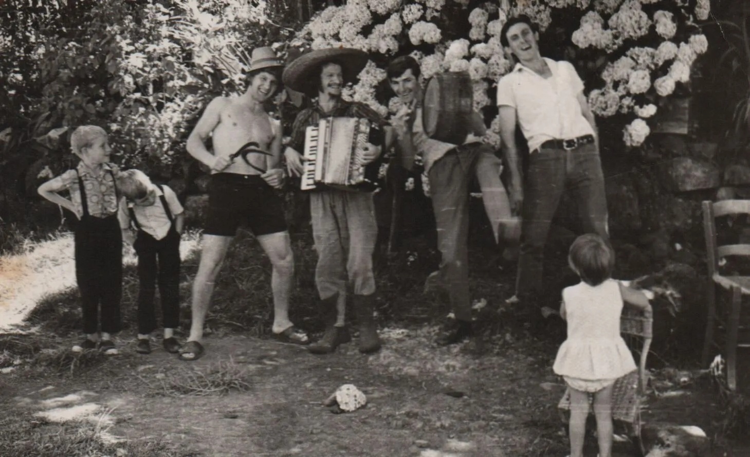 Um grupo de sete homens, jovens e crianças, em pé, em um ambiente externo com flores brancas e folhagem ao fundo, um homem está tocando acordeão e usando um chapéu mexicano enquanto os outros riem. (Original)