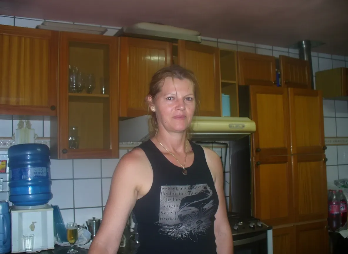 Mulher de meia-idade de cabelos loiros e ombros largos, em uma blusa preta, sorri para a câmera em uma cozinha com armários de madeira e azulejos brancos.