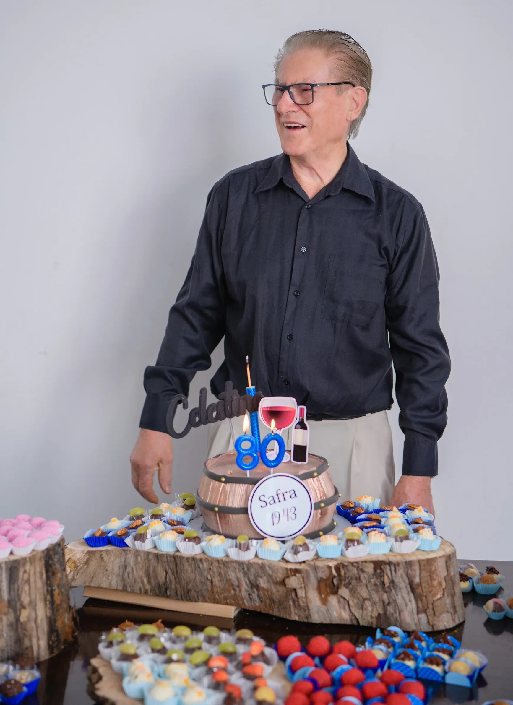 Homem de óculos, camisa social escura e calças claras, sorrindo e olhando para o lado direito, em pé atrás de uma mesa com um bolo de aniversário decorado com o número "80", uma garrafa de vinho, uma taça e docinhos variados.