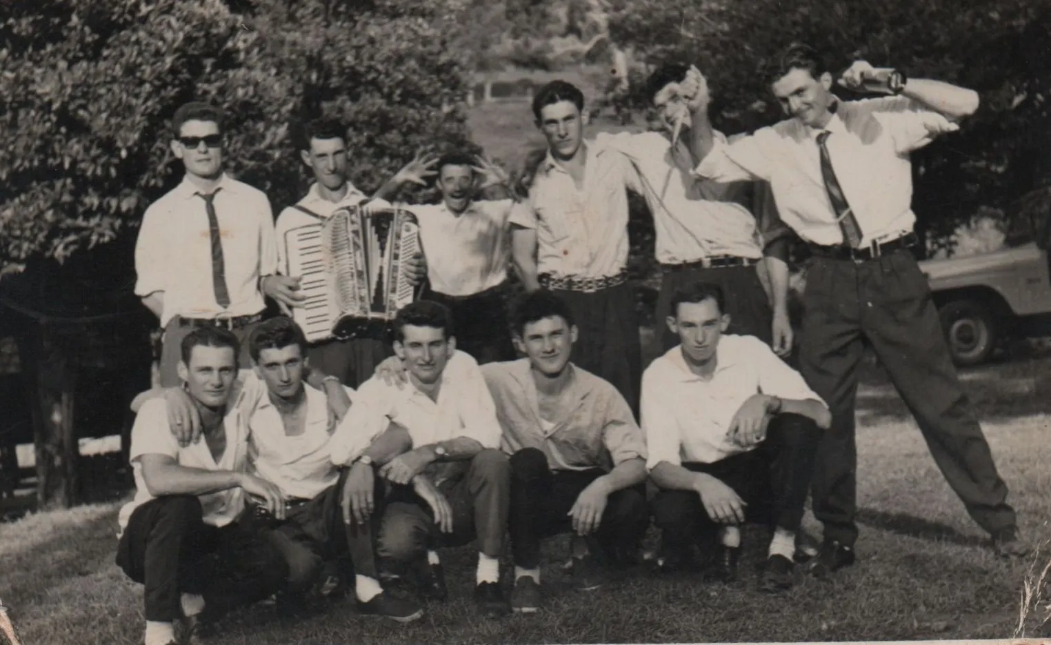 Um grupo de dez jovens posa ao ar livre. Um dos rapazes senta-se no centro a tocar um acordeão, enquanto os outros estão sentados ou em pé à sua volta, a sorrir. (Original)