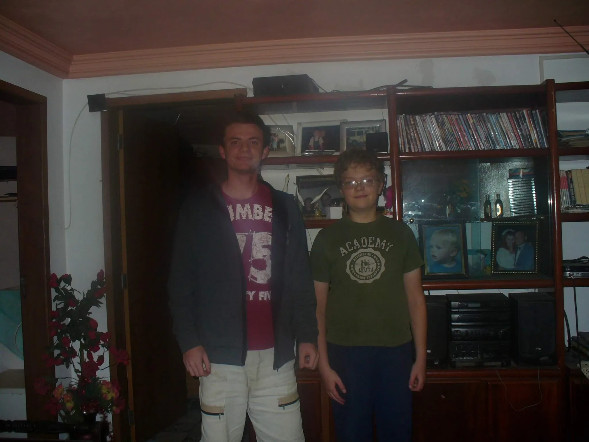 Dois homens, um de estatura mais alta, em pé dentro de uma casa, próximos a uma estante cheia de livros e CDs.