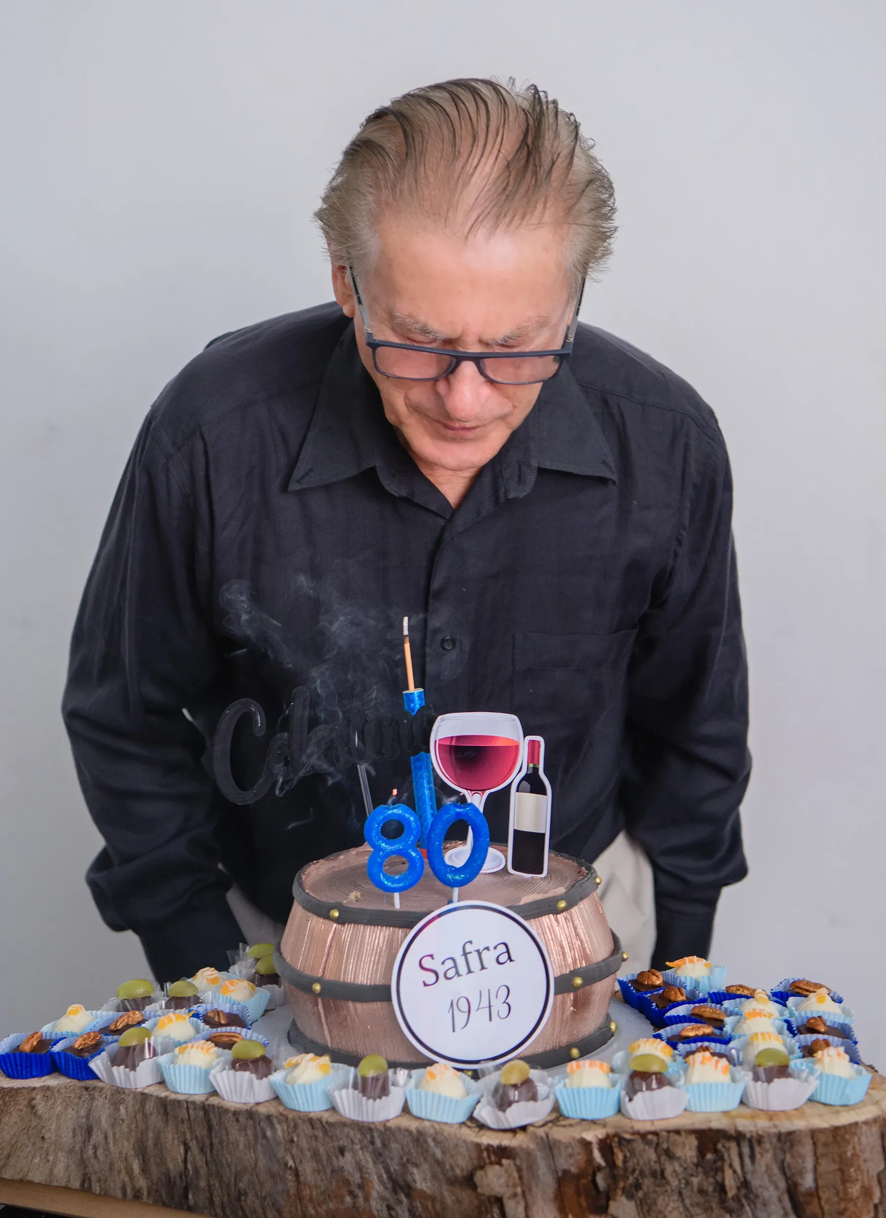 Um homem de óculos, vestindo uma camisa escura, sopra uma vela de aniversário azul com o número 80 em um bolo em forma de barril, cercado por doces em uma tábua de madeira.