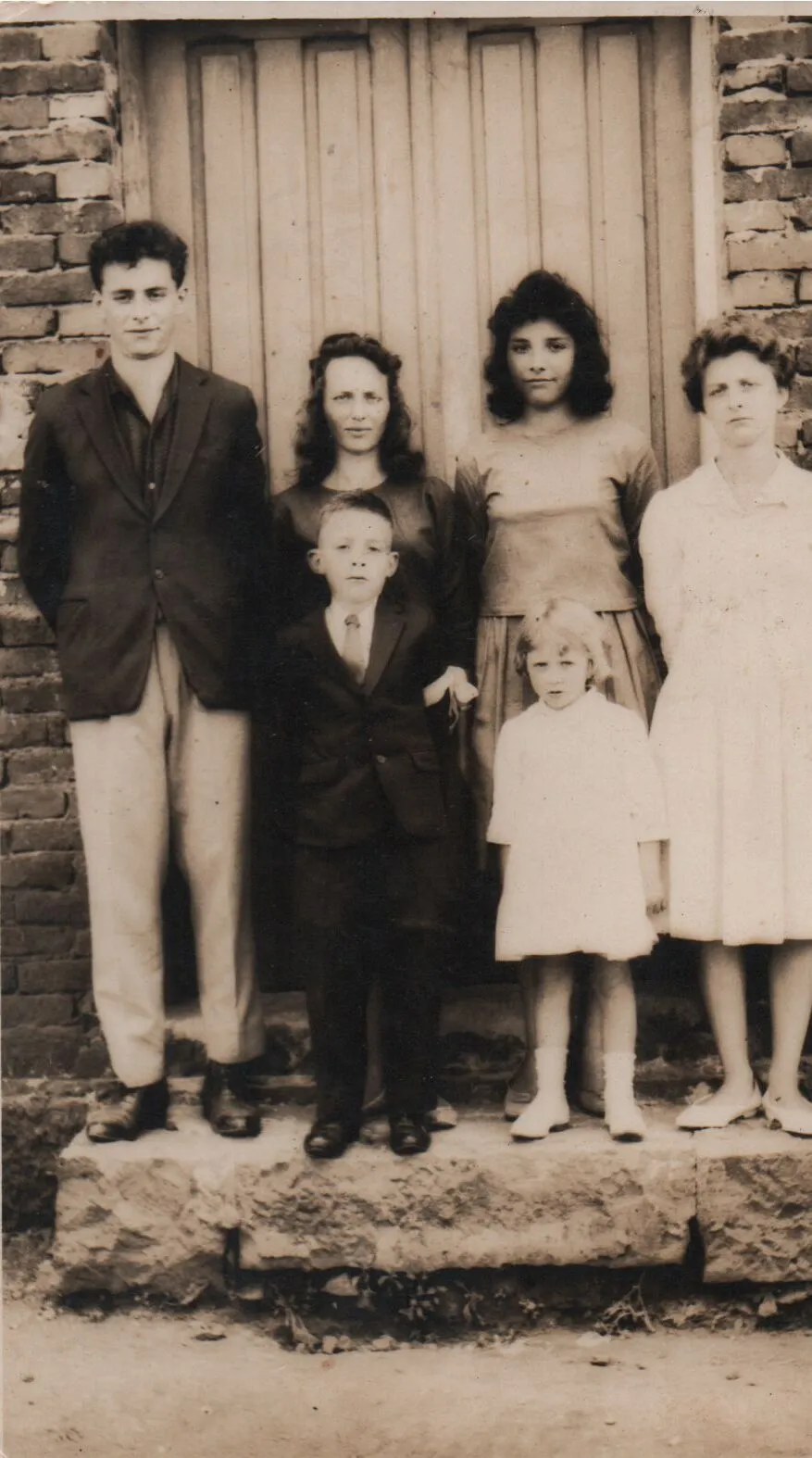 Grupo de cinco pessoas, três mulheres e dois meninos, posam em frente a uma porta azul-esverdeada com uma parede de tijolos vermelhos de cada lado. O menino do centro e o homem mais velho da esquerda estão vestidos com ternos pretos, enquanto as mulheres e a menina da direita estão vestidas com vestidos brancos e azuis. (Original)