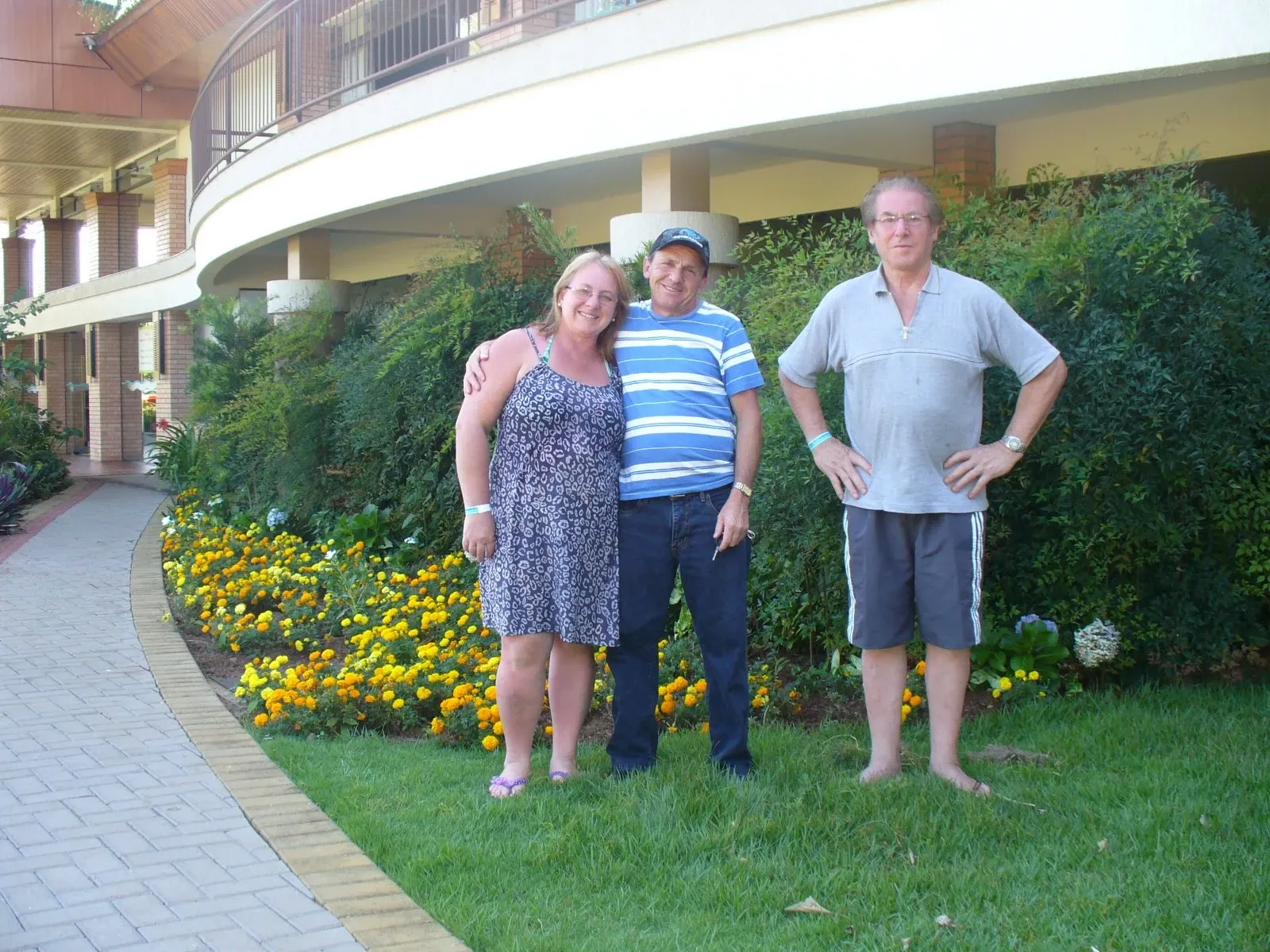 Três indivíduos, um homem, uma mulher e um segundo homem, estão em pé na grama verde ao lado de um canteiro de flores.