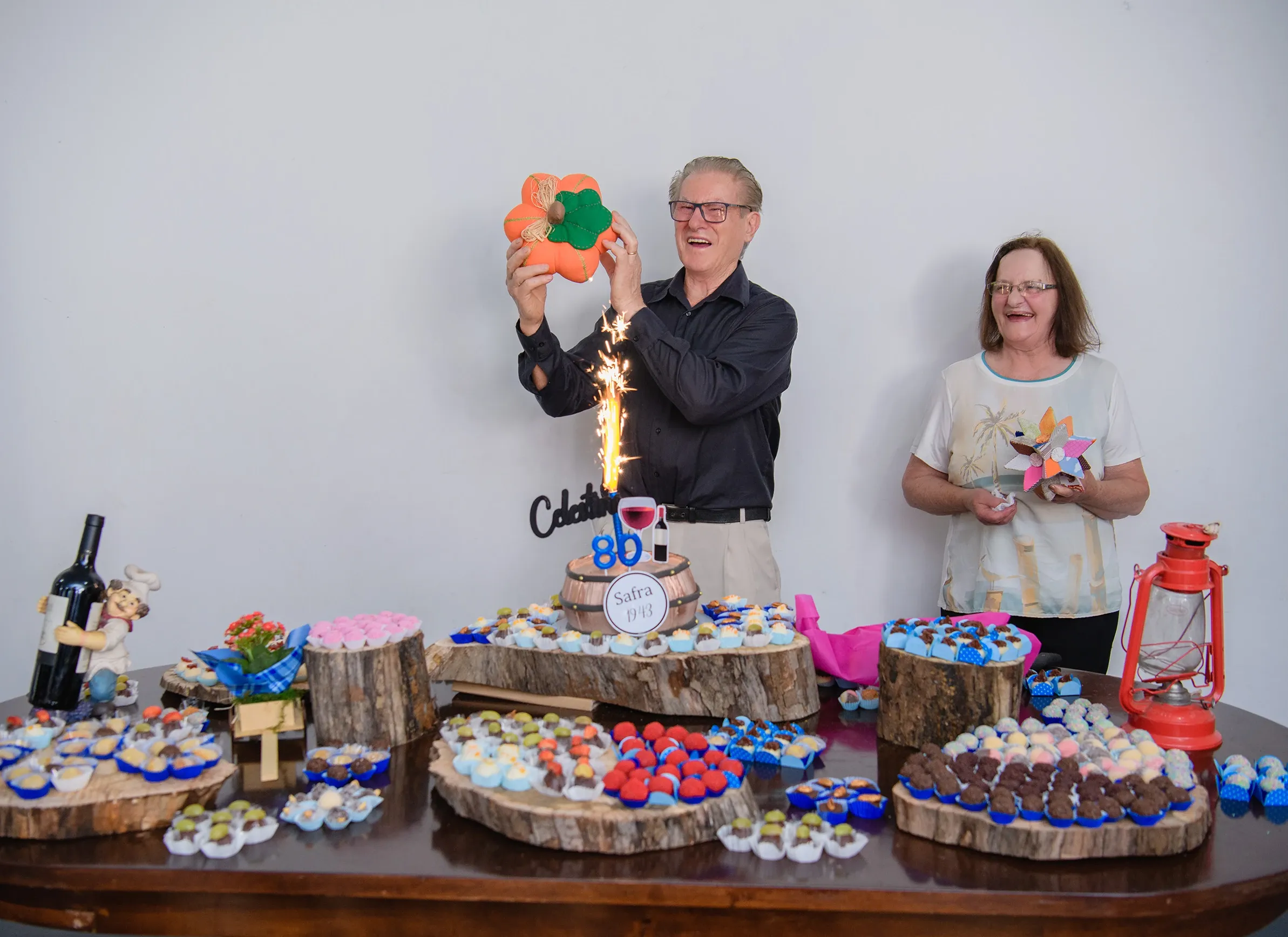 Um homem e uma mulher rindo e celebrando em uma festa de aniversário de 80 anos com muitas guloseimas em uma mesa de madeira. O homem segura uma abóbora decorativa e um pavio luminoso solta faíscas perto do bolo de aniversário.