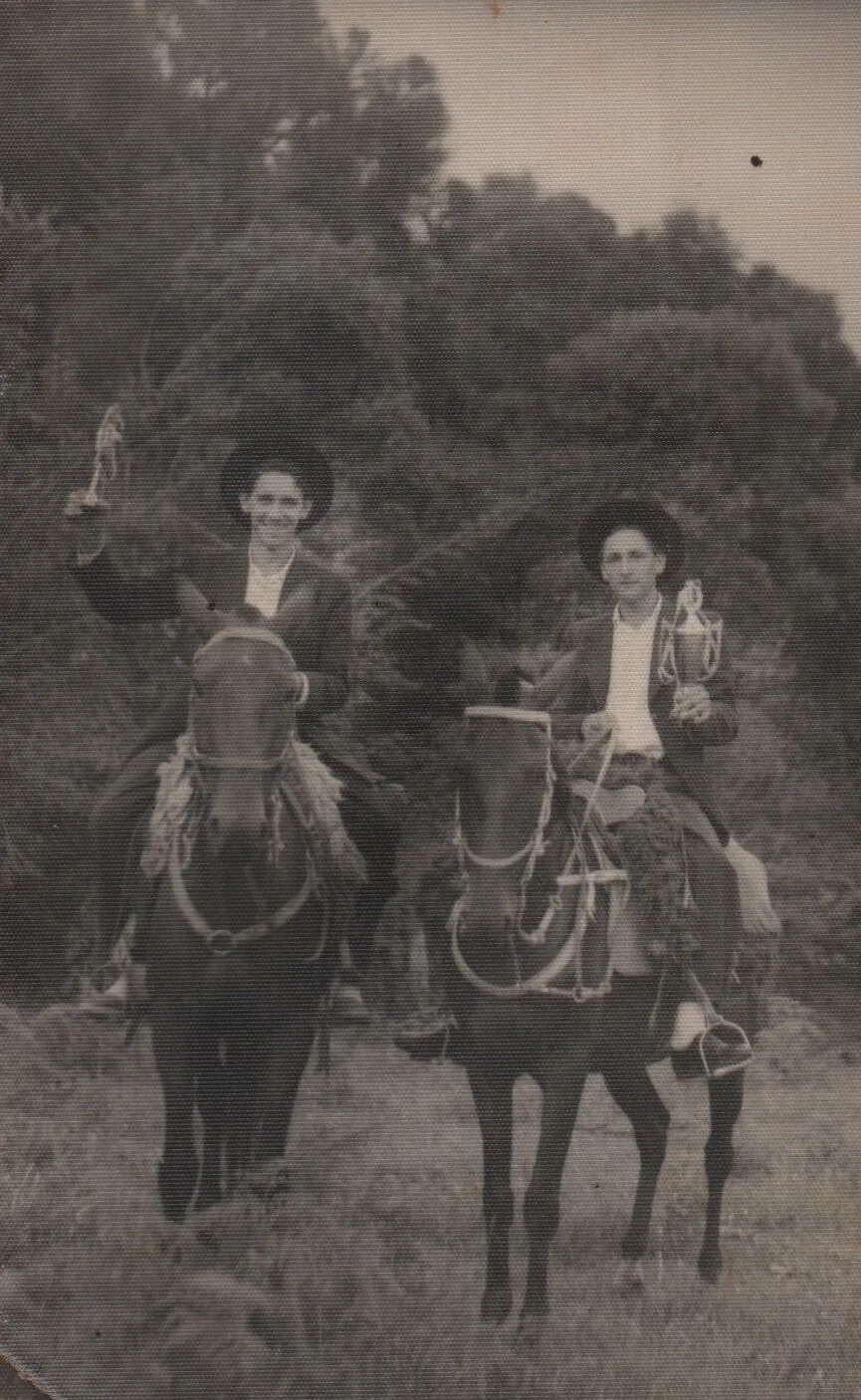 Dois homens sorridentes, vestidos com trajes formais escuros e chapéus marrons, montam a cavalo em uma área gramada com árvores ao fundo, cada um segurando um troféu dourado. (Original)