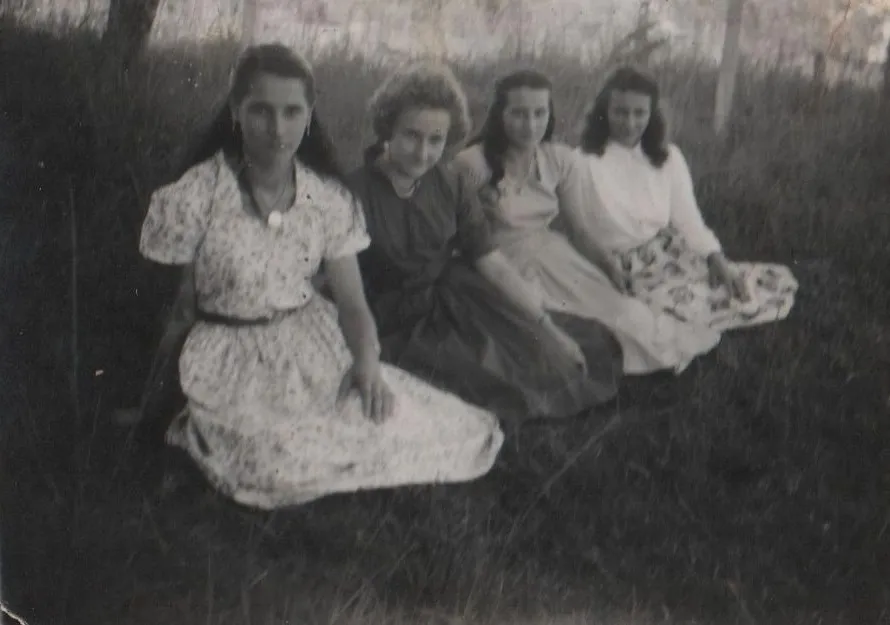 Quatro mulheres jovens com diferentes penteados e cores de cabelo estão sentadas na grama, vestindo vestidos florais de verão. (Original)