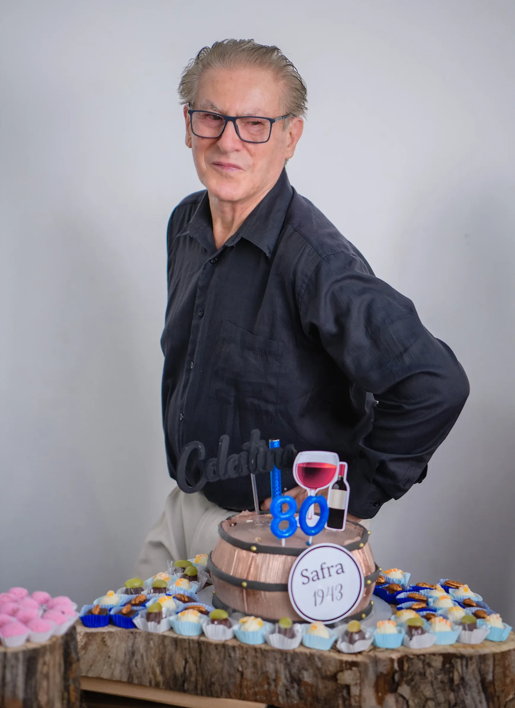 Homem idoso de óculos e camisa preta, sorrindo e com as mãos nos quadris, em pé atrás de um bolo de aniversário em formato de barril de vinho e pequenos doces em uma mesa de madeira.