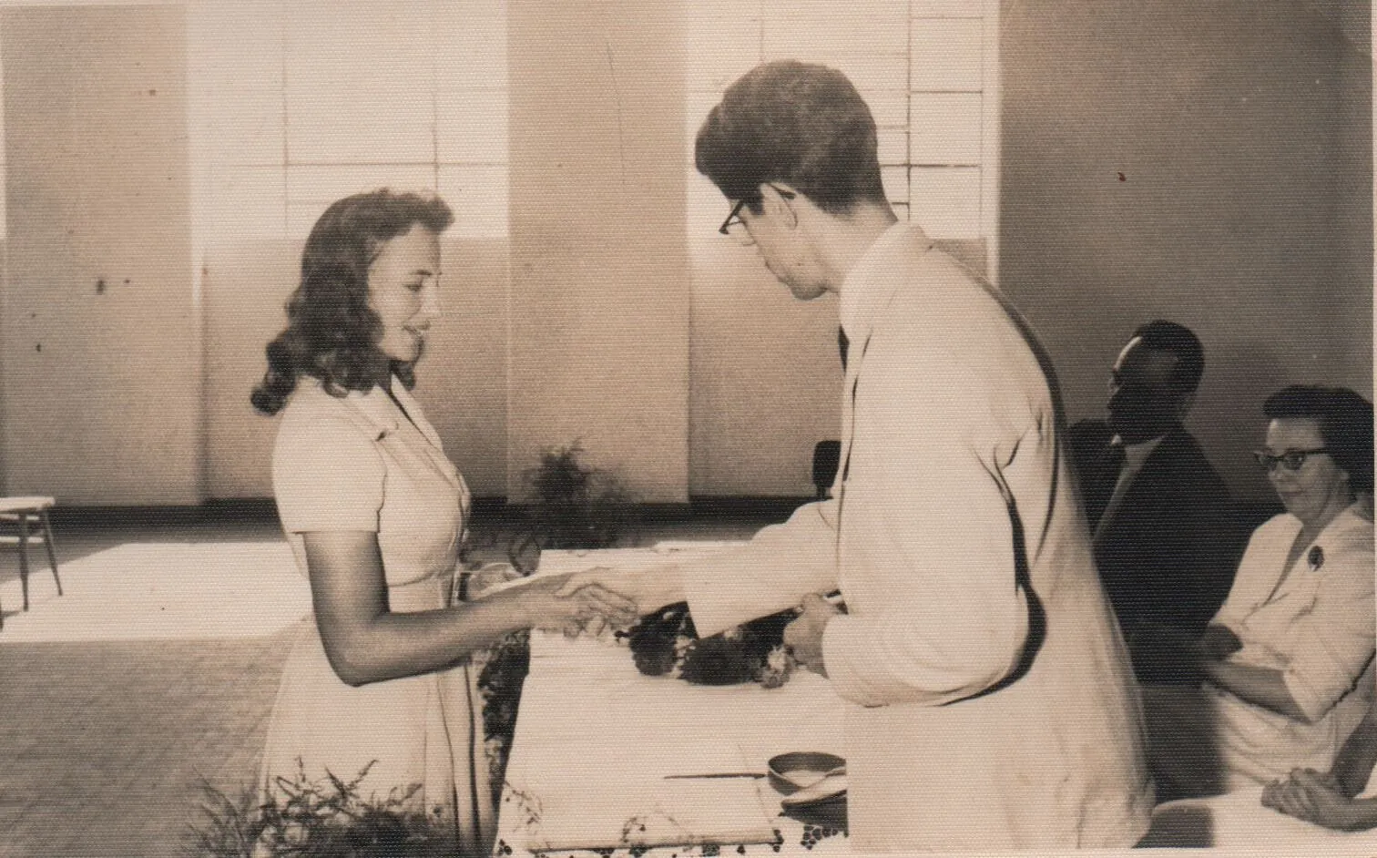 Uma jovem de vestido azul se inclina e aperta a mão de um homem de terno bege, que sorri para ela. (Original)
